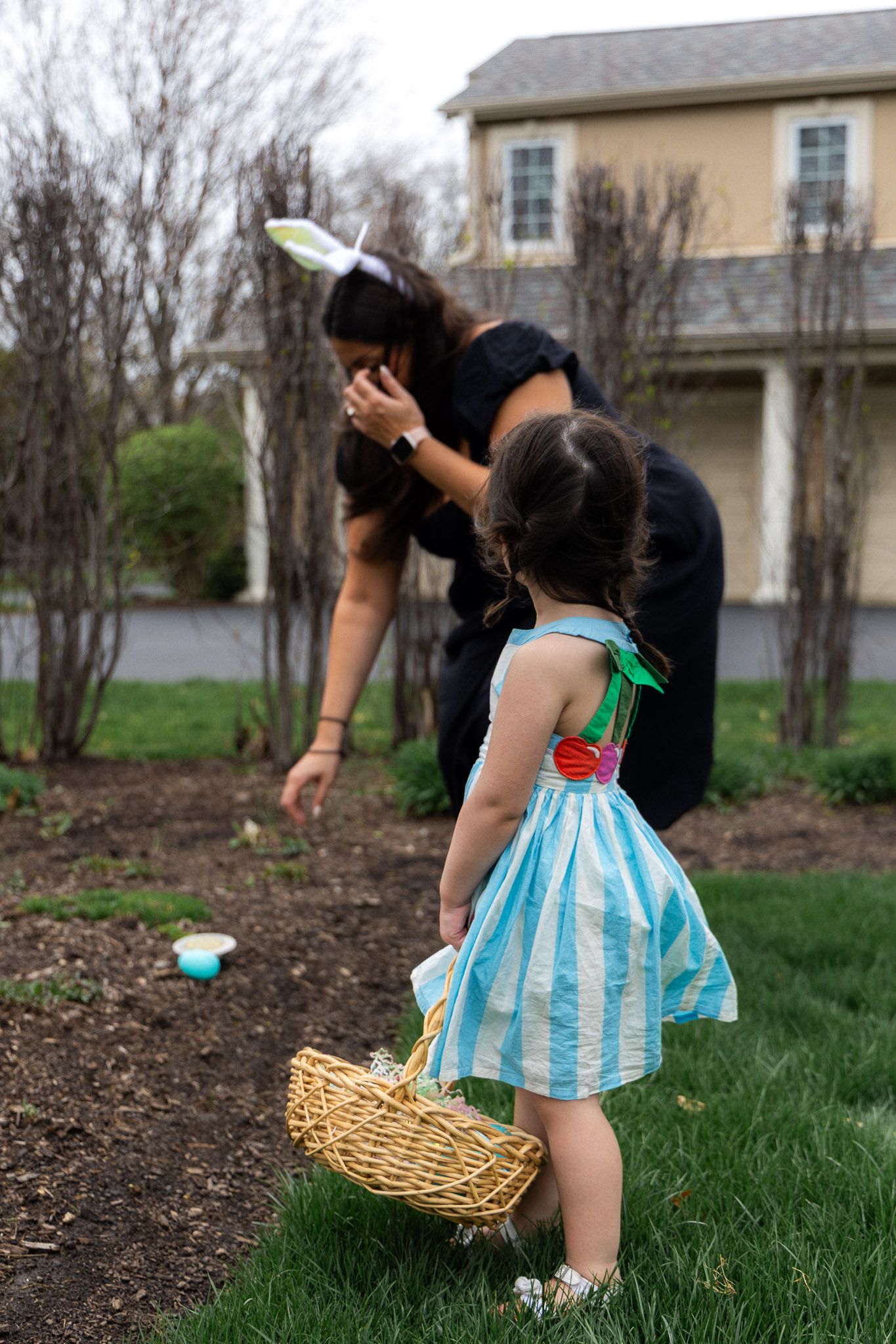 Girl in blue dress holding Easter basket watches woman in bunny ears place eggs in garden.