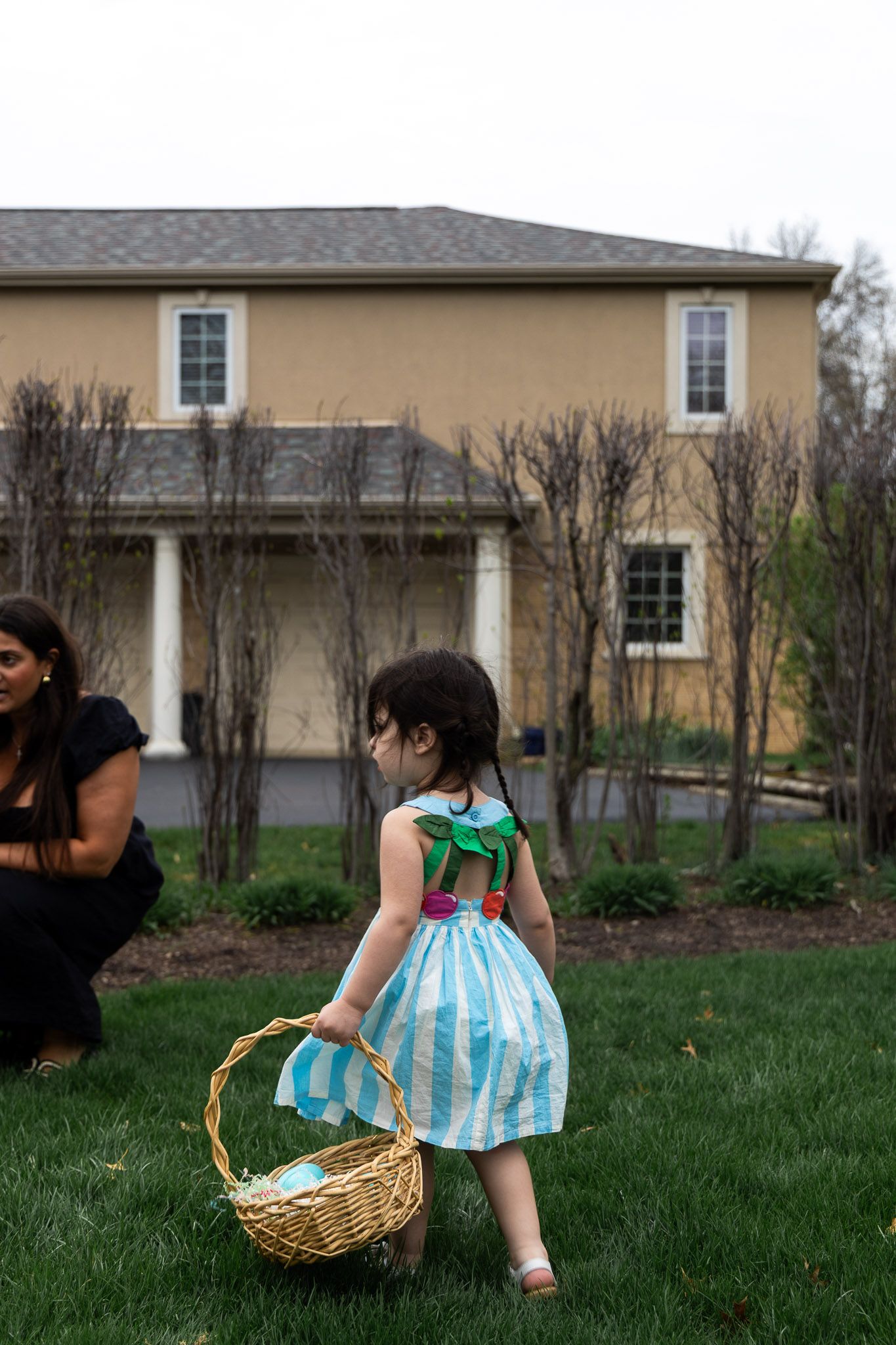 A young girl in a blue striped dress carries a wicker Easter basket across a green lawn in front of a large home.
