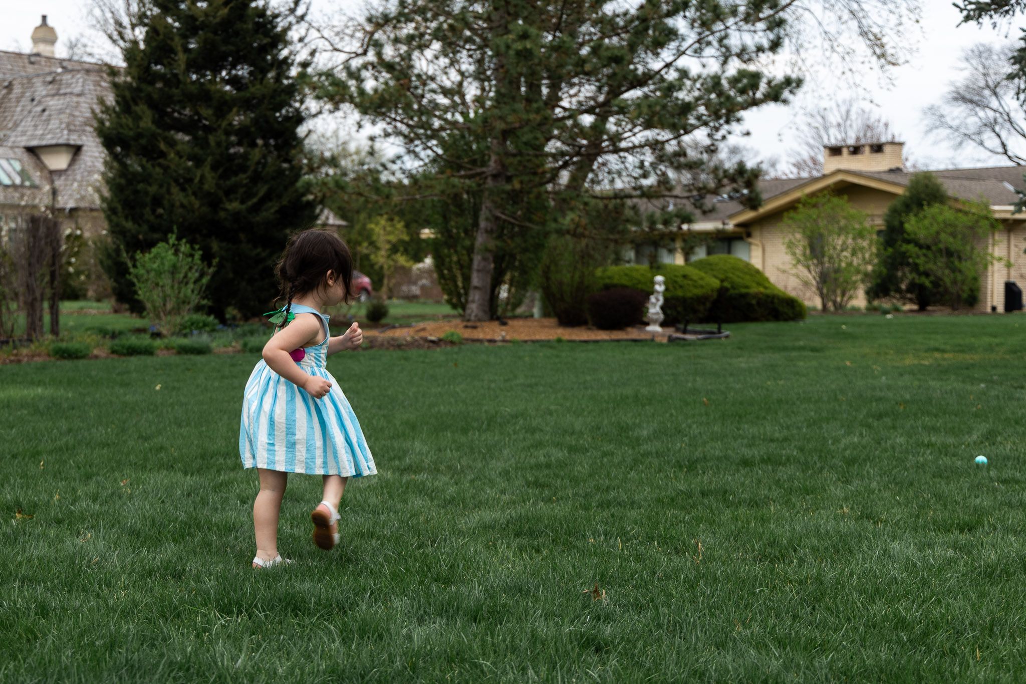 A little girl in a blue striped dress runs barefoot across a lush green lawn during an Easter egg hunt.