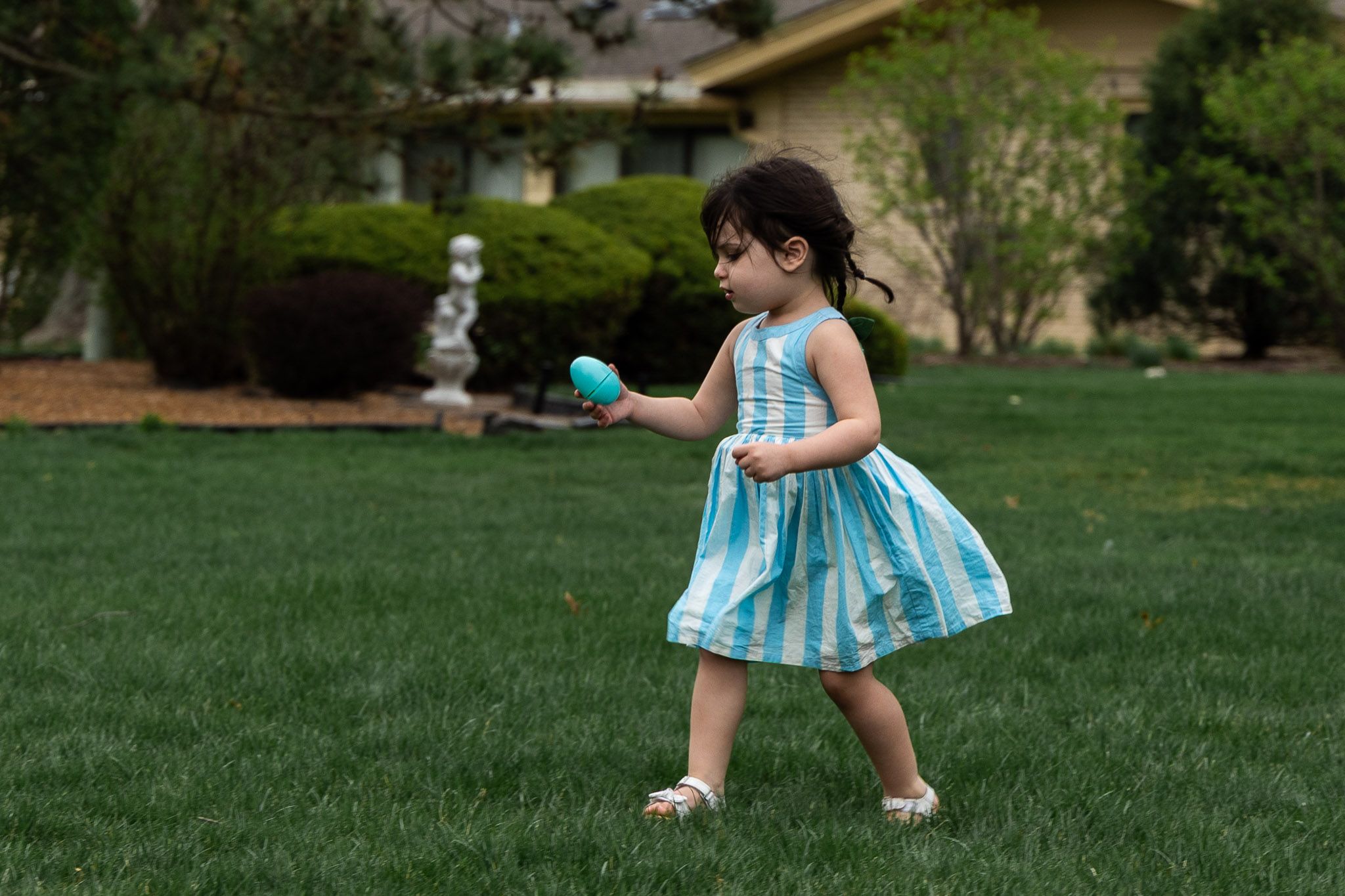 Toddler girl in blue striped dress running on green grass holding a teal Easter egg during an outdoor egg hunt.