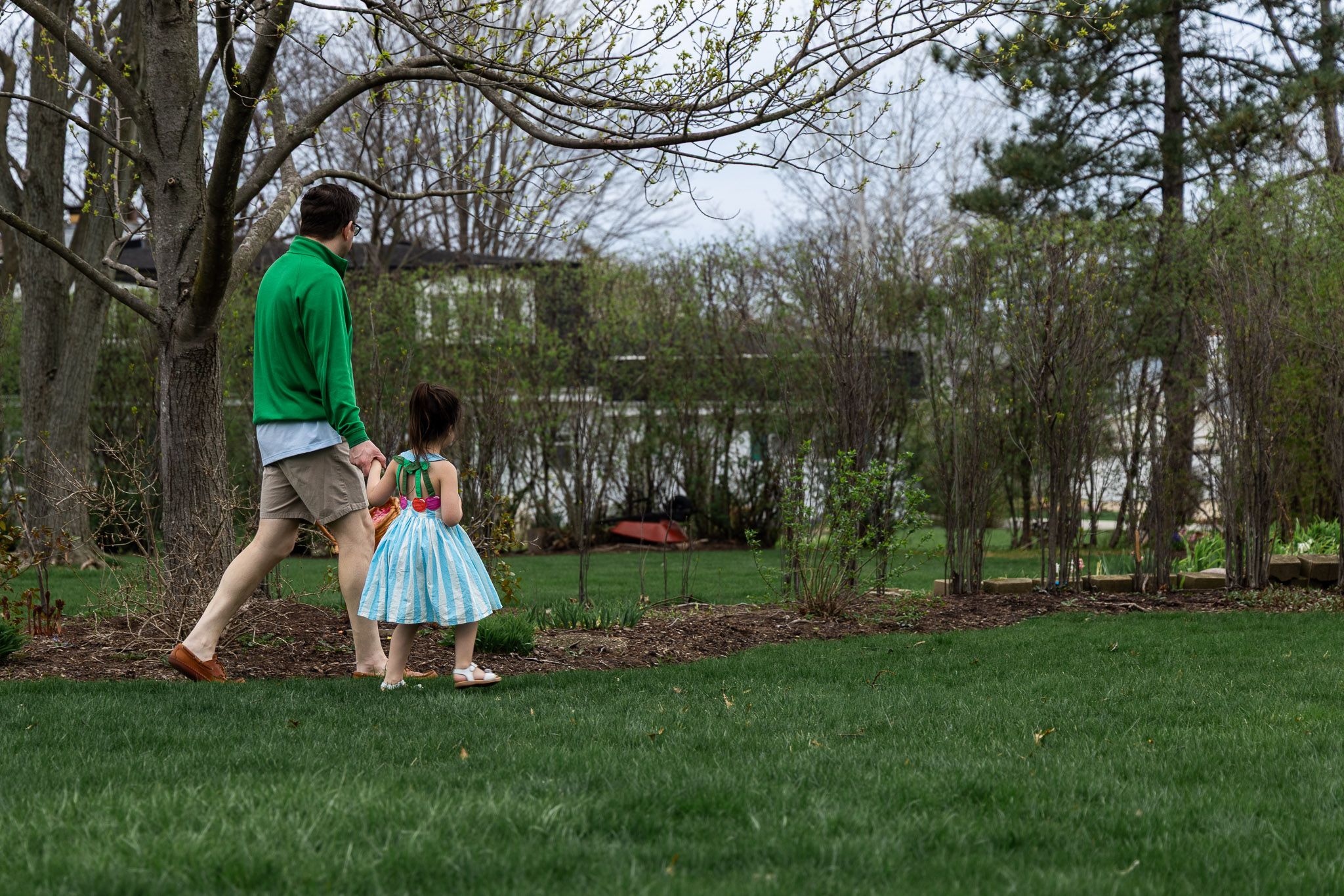 A father and young daughter walk hand-in-hand through a lush green yard near a pond surrounded by trees.