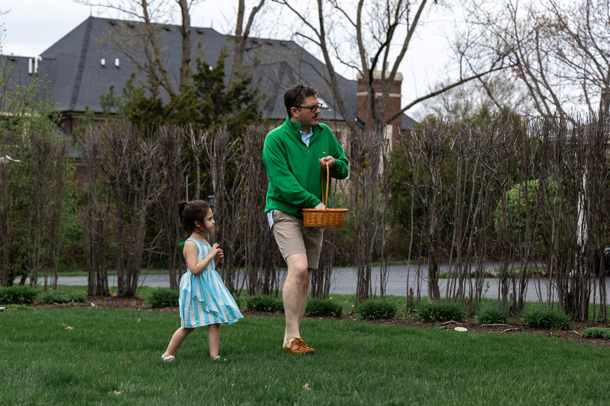 Man in green hoodie carrying Easter basket with young girl in blue dress during outdoor Easter egg hunt.
