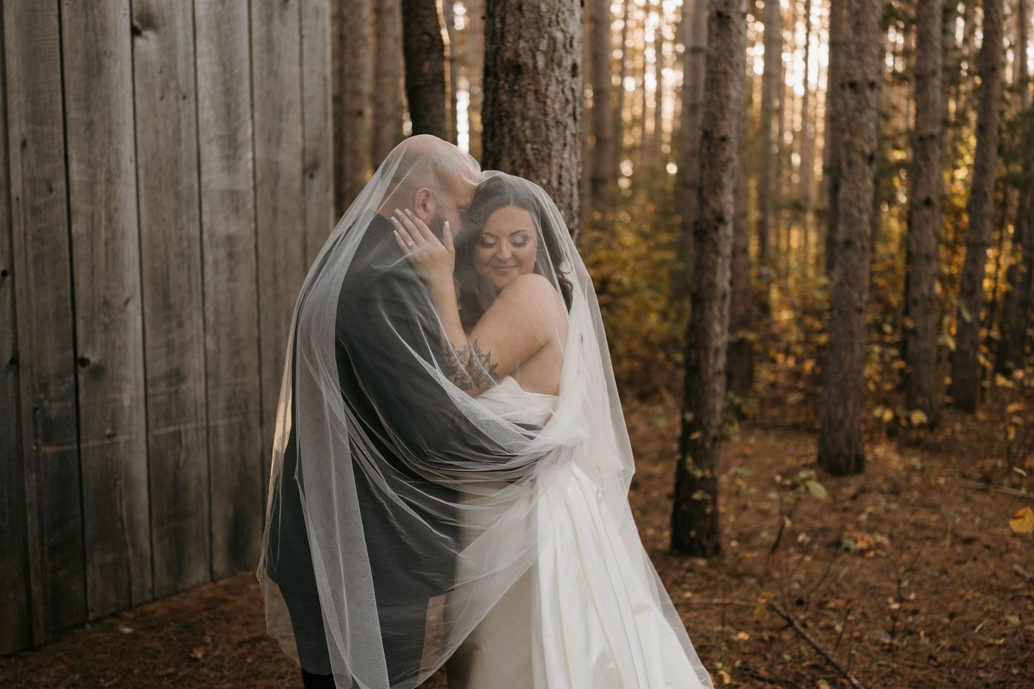 Bride and groom sharing a romantic moment under a flowing veil in an autumn forest.