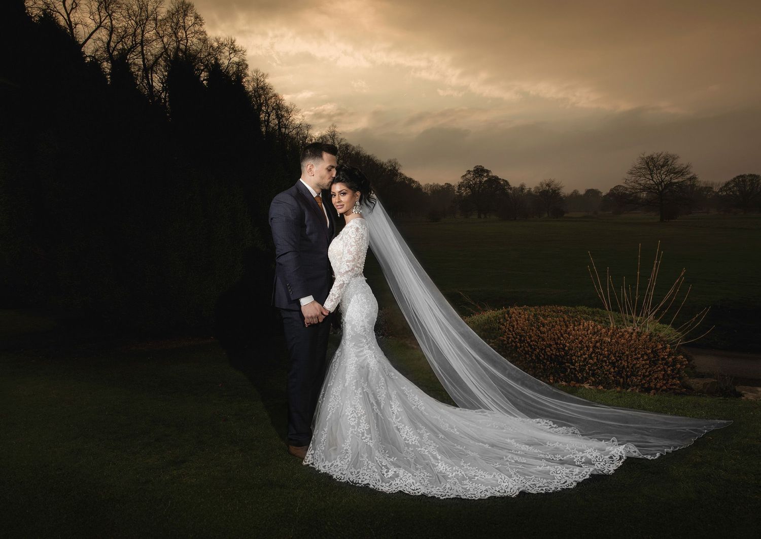 Bride and groom cutting their white tiered wedding cake on a wooden stand at a romantic, dimly lit wedding reception.