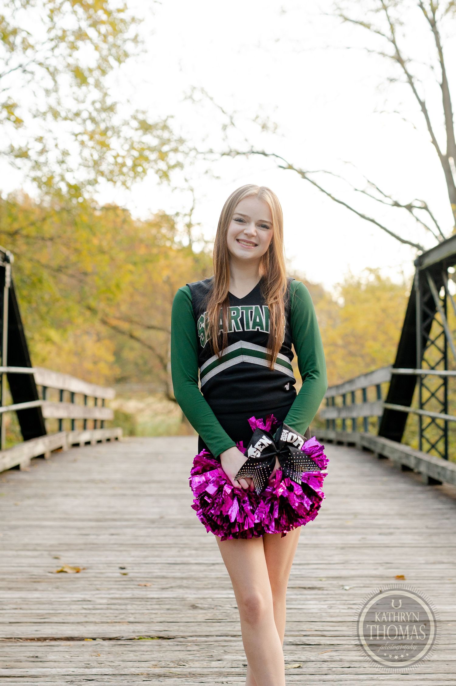 Cheerleader smiles while holding bright pink pom poms on rustic bridge with fall foliage in background.