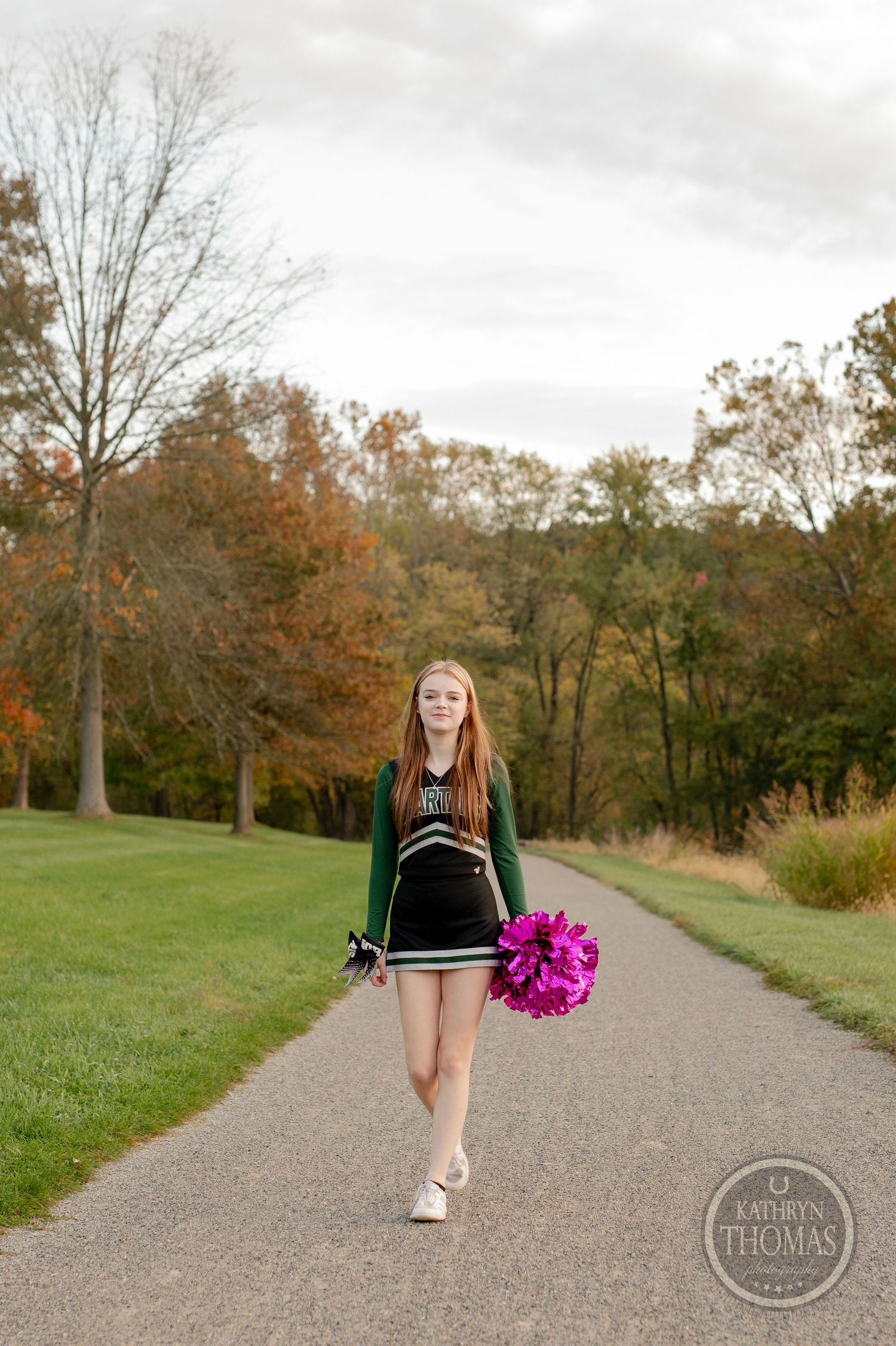Cheerleader in green and black uniform walks path holding bright pink pom pom.