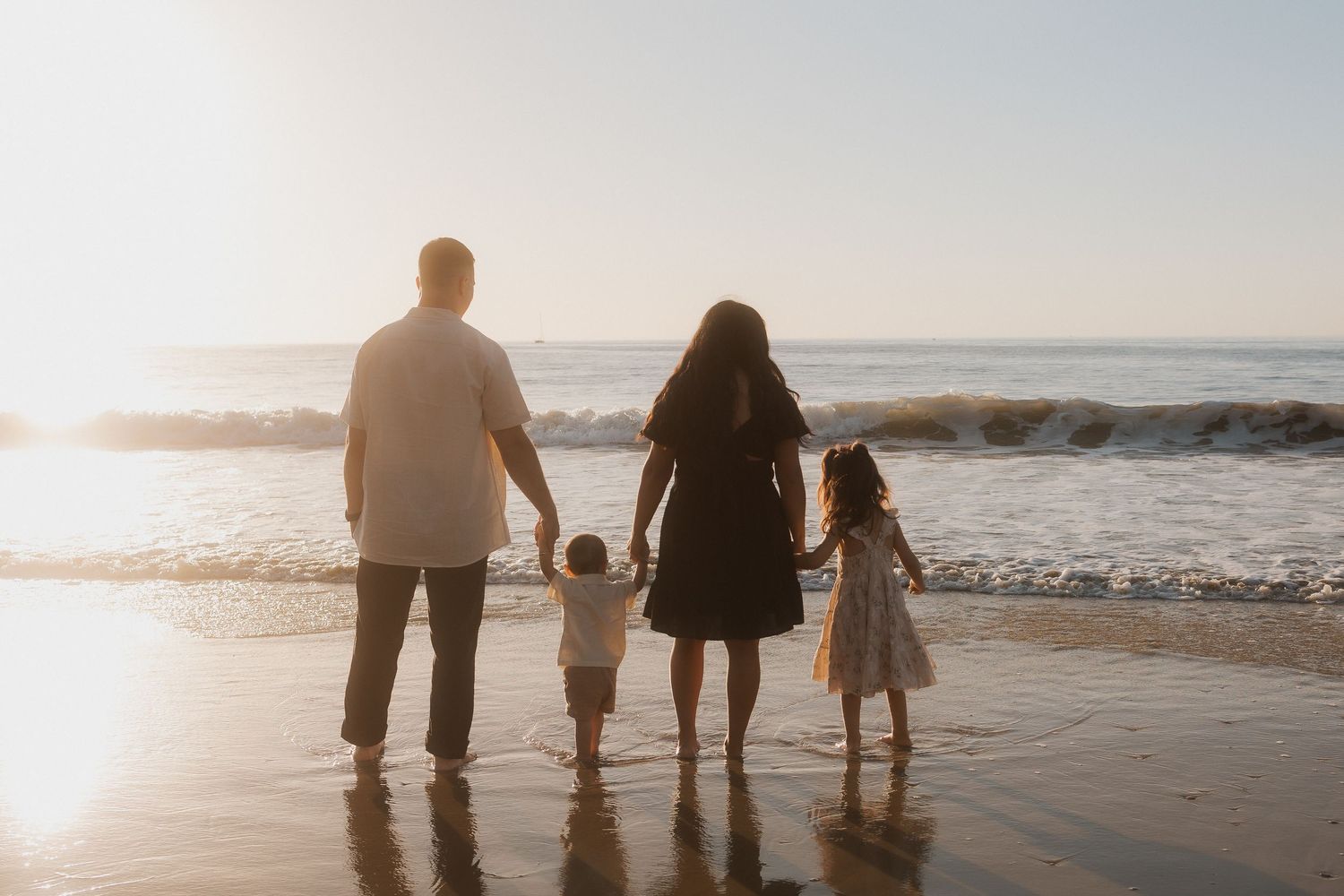 Silhouetted family holding hands while walking on beach at sunset beside ocean waves.