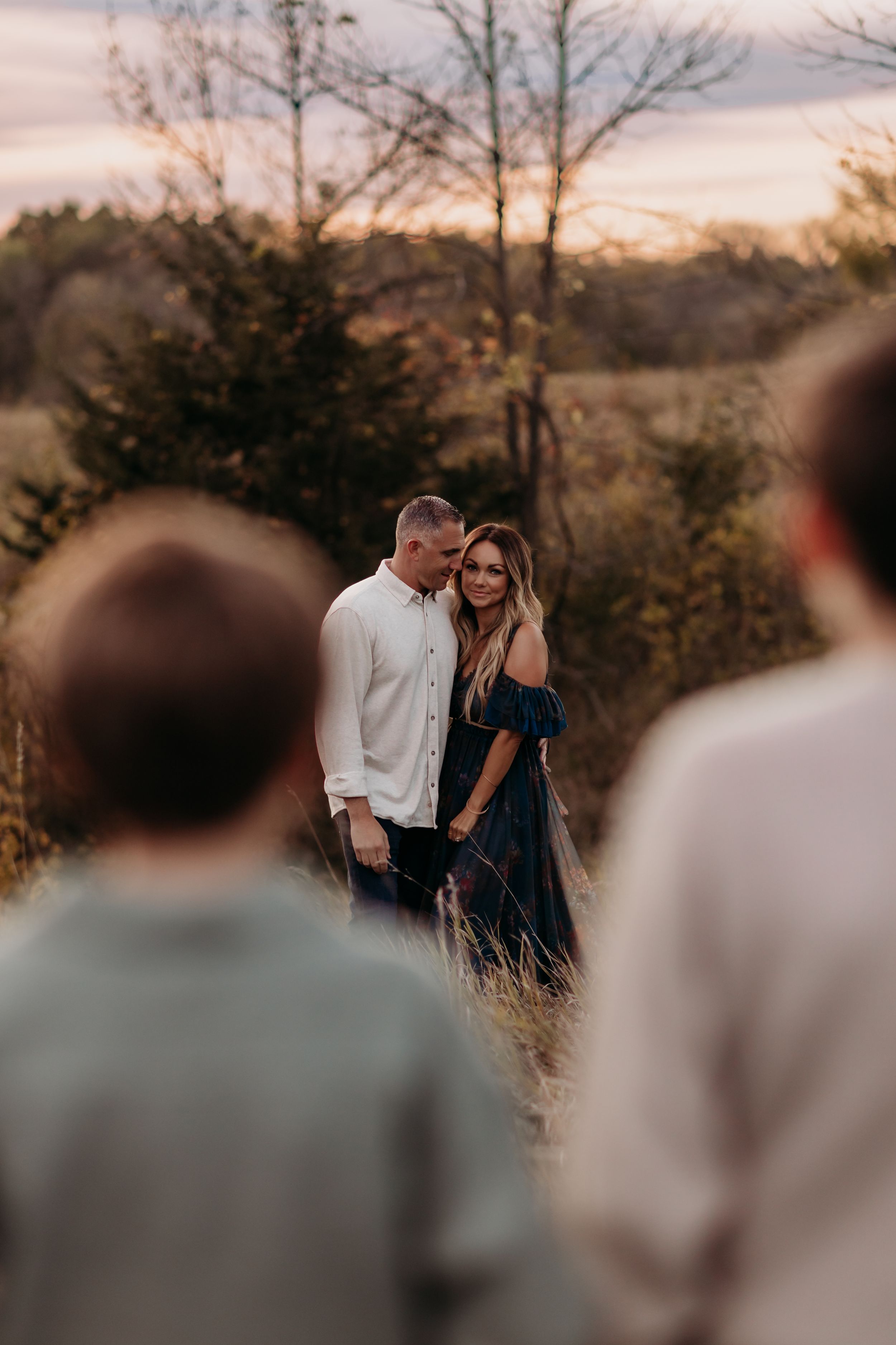 Couple embracing at golden hour in a field, framed by blurred figures in foreground, surrounded by autumn trees.