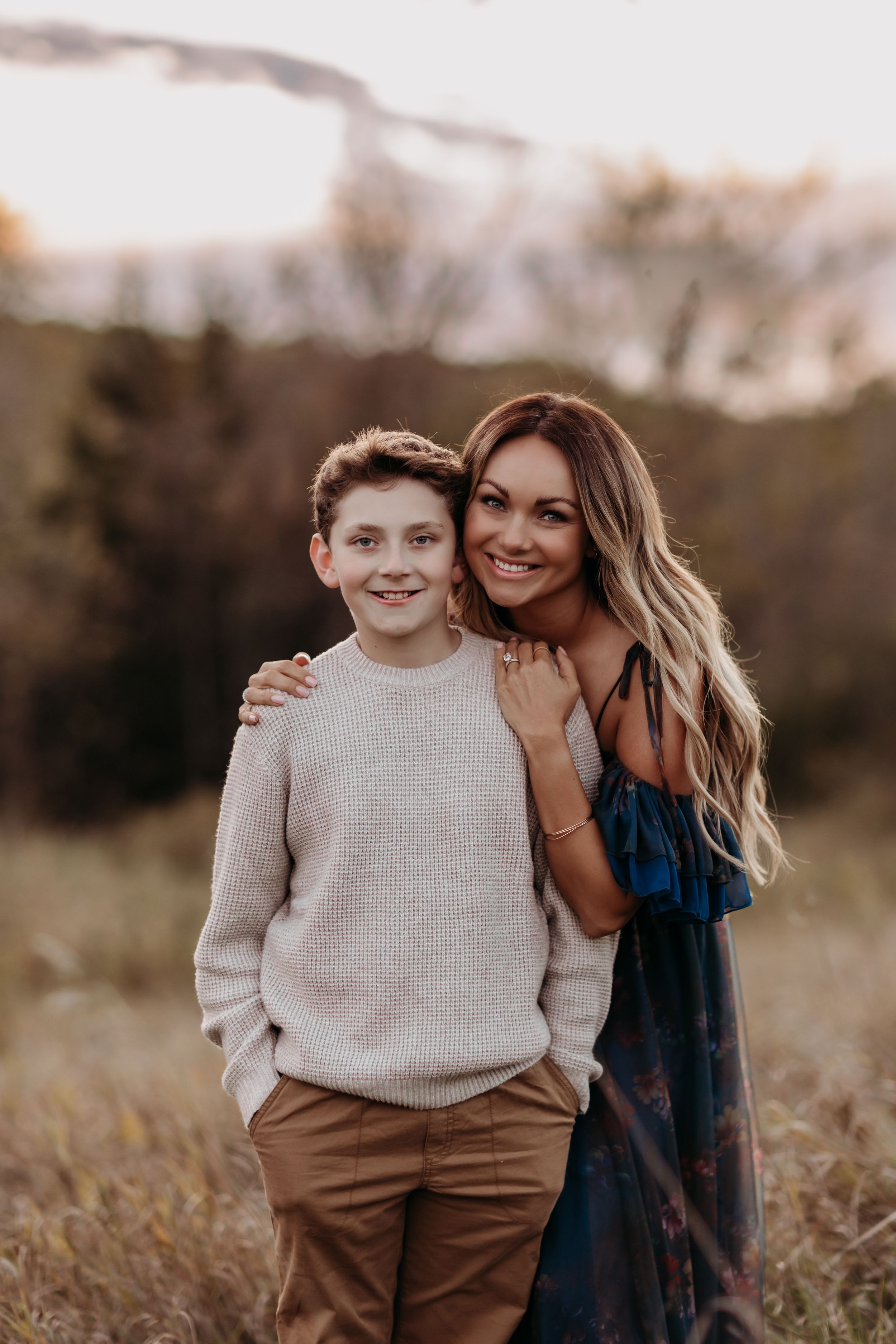 A smiling woman in a blue floral dress poses with a young boy in a beige sweater outdoors at golden hour.