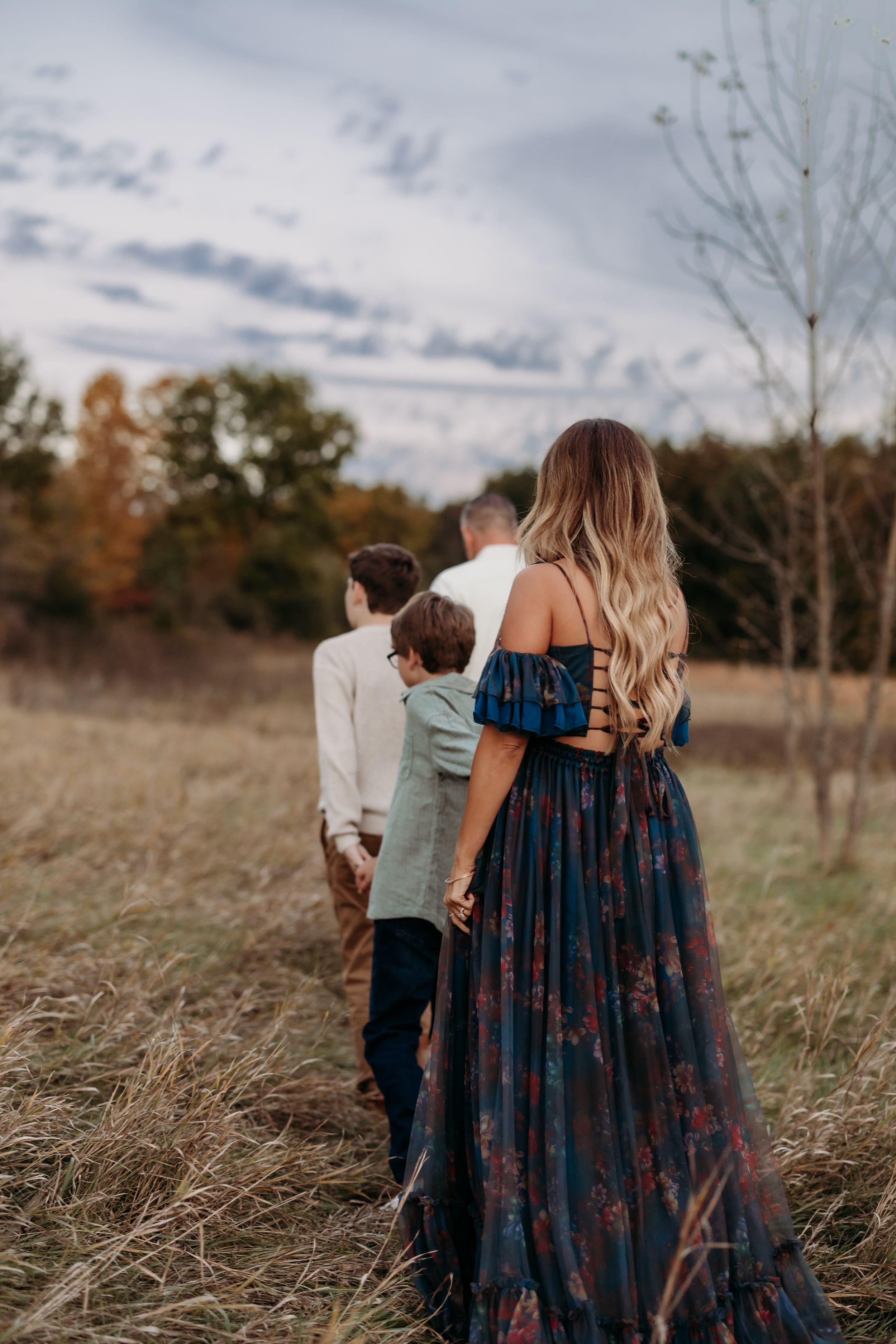 Family walking through autumn field, woman in floral off-shoulder gown with two boys and man heading toward fall foliage.