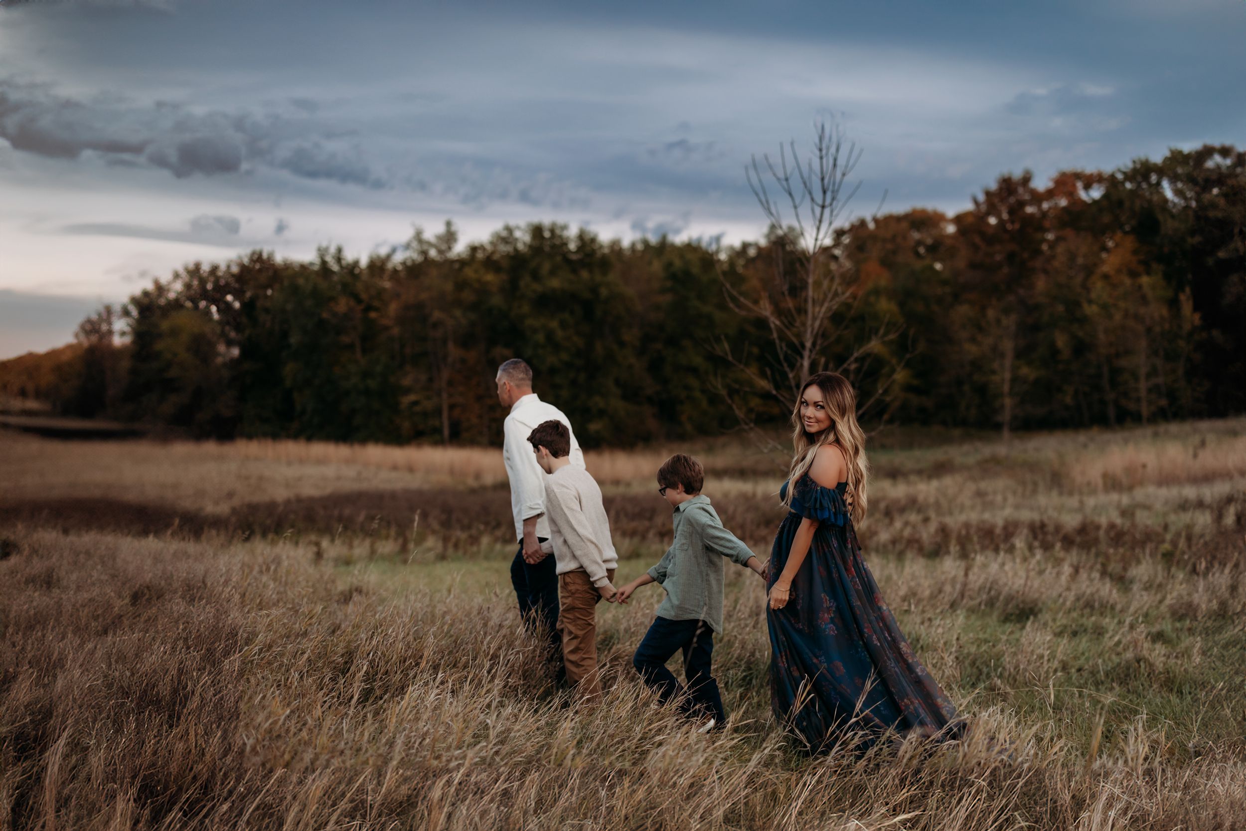 Family of four walking through autumn field at dusk, holding hands, woman in flowing navy gown.