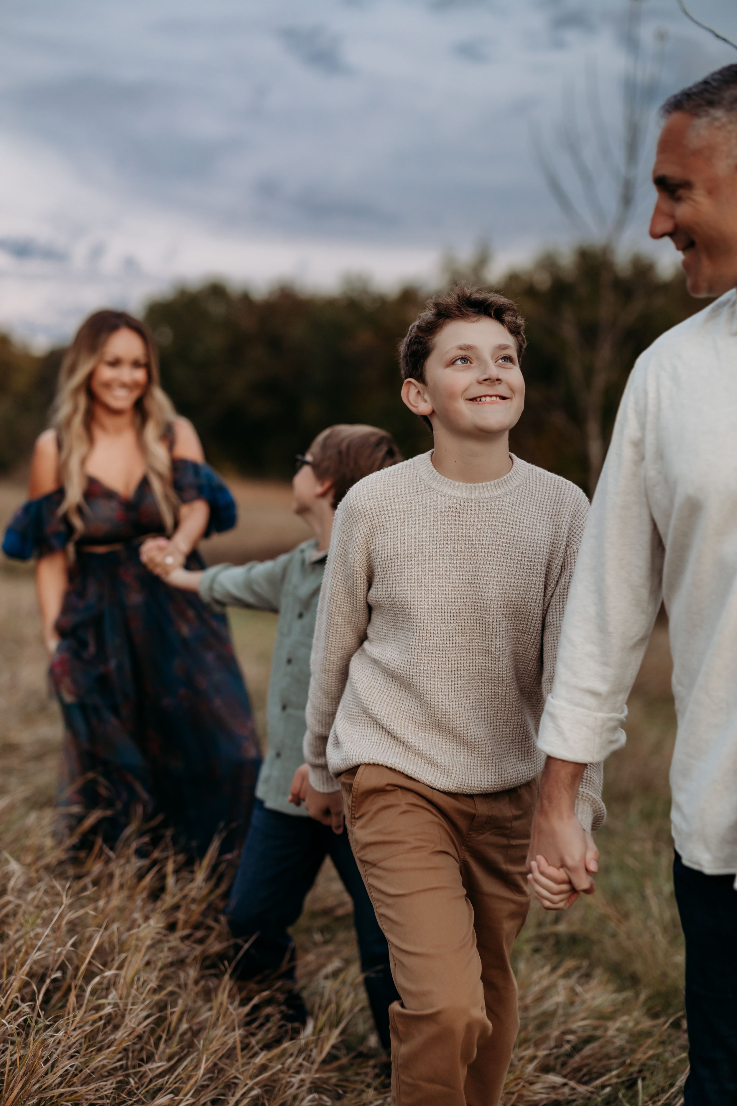 A smiling boy in a beige sweater walks through a field with his family during an outdoor photo session at dusk.