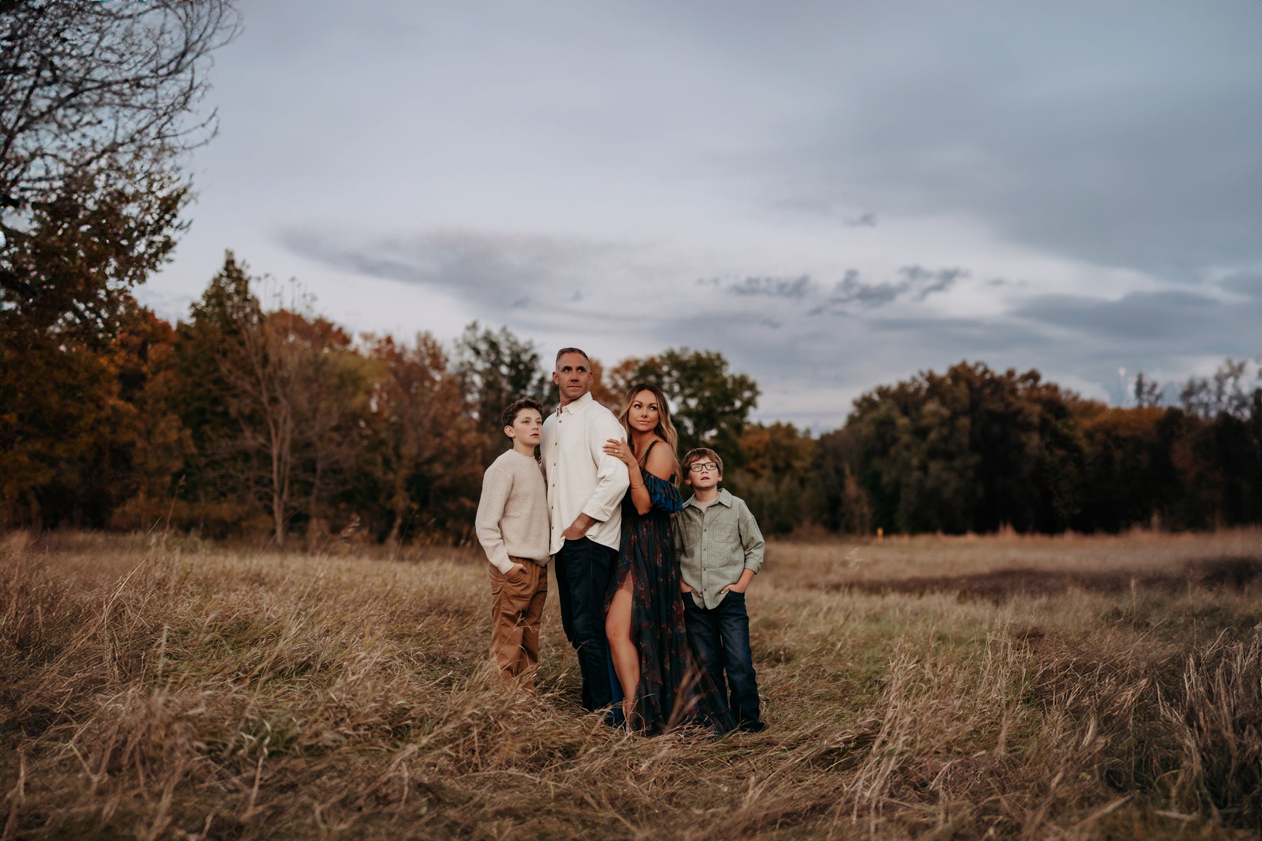 Family of five posing together in an autumn field with colorful trees and overcast sky.