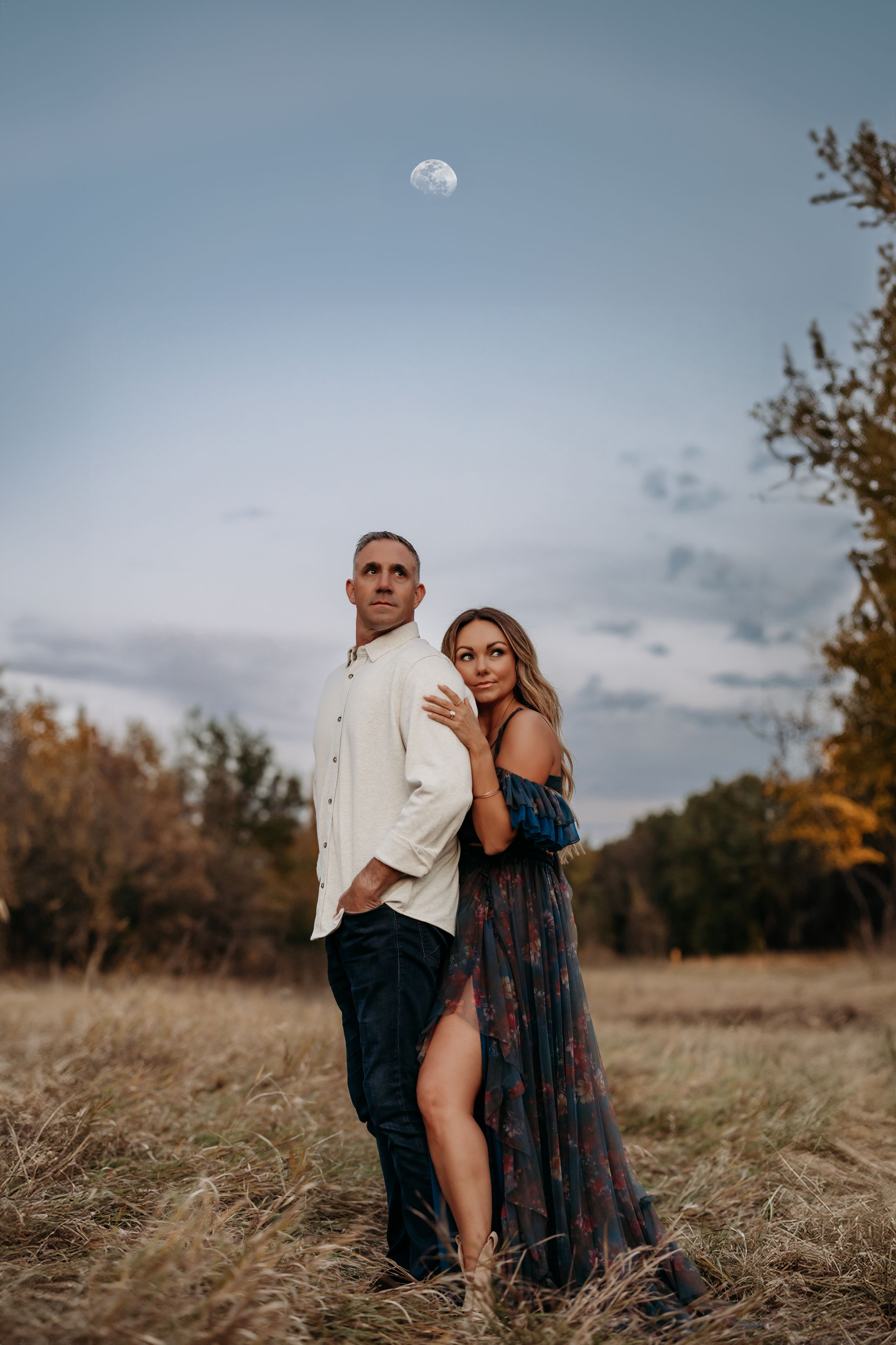 Couple posing in a golden field at dusk with a moon overhead, woman in floral gown embracing man in white shirt.
