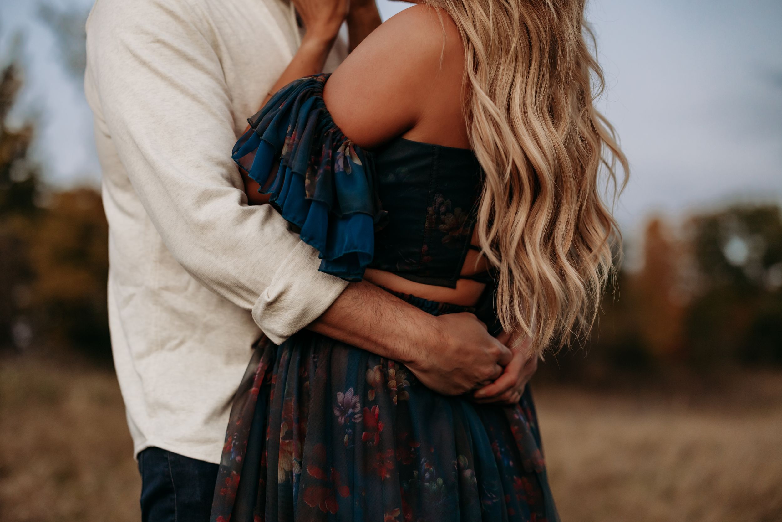 Couple embracing outdoors in autumn, woman in floral off-shoulder dress with long blonde hair, man in white shirt.