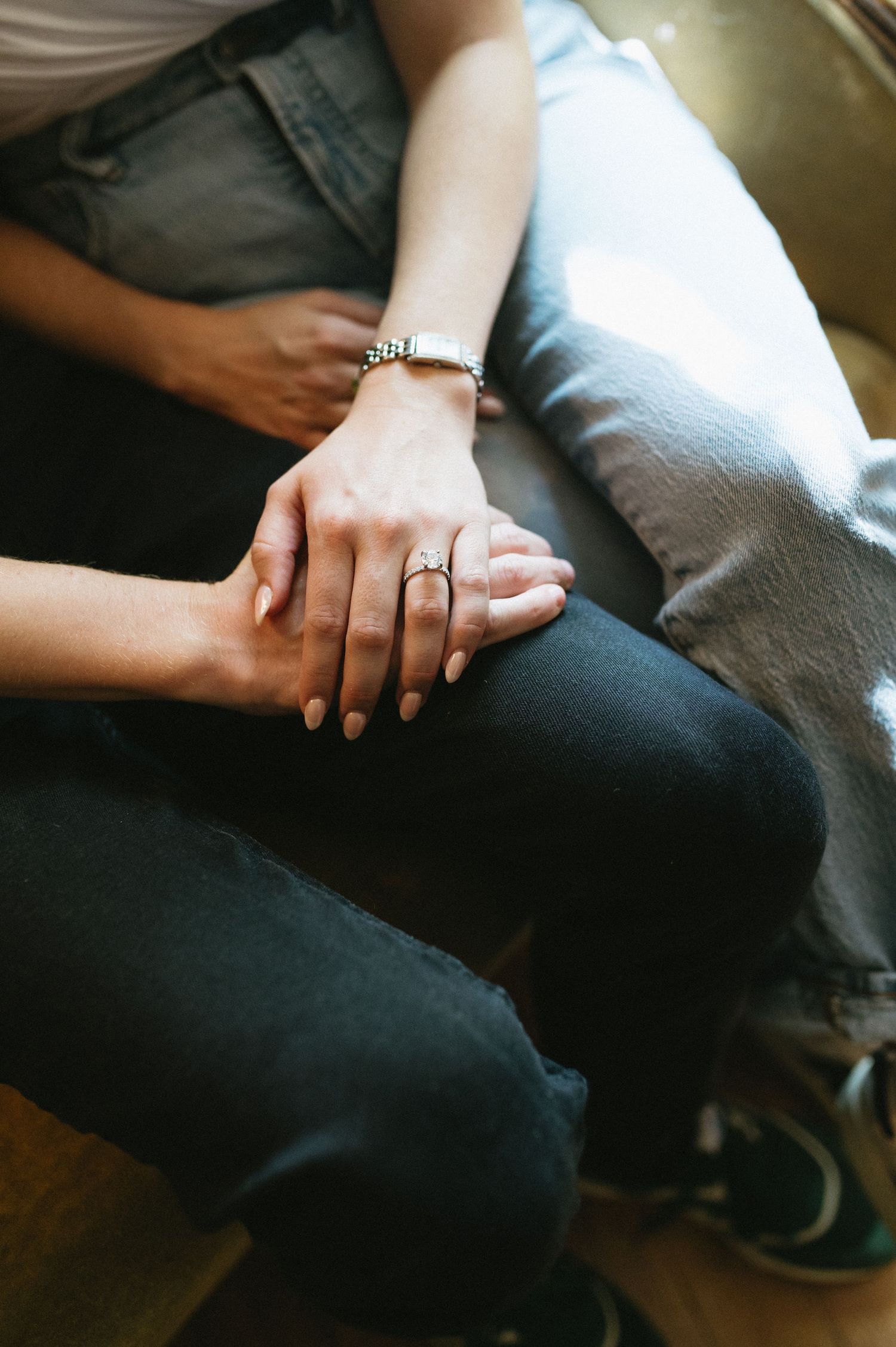 Intimate close-up of intertwined hands with jewelry resting on denim-clad legs.