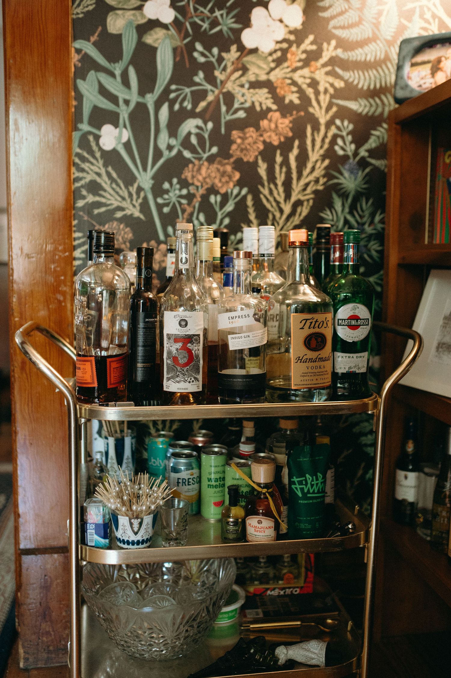 Well-stocked vintage bar cart with various liquor bottles against floral wallpaper.