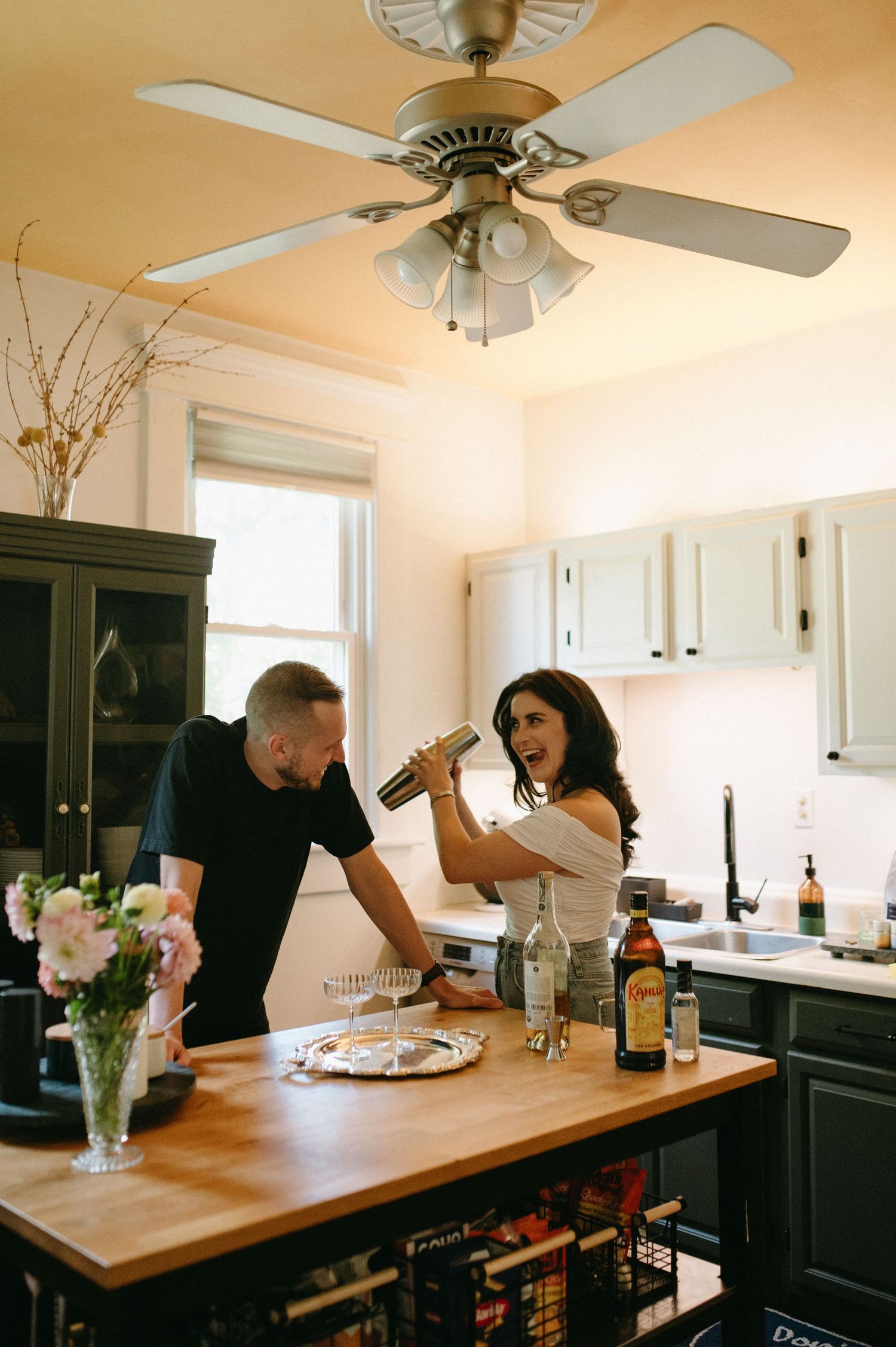 Two people share drinks in a cozy kitchen with dark cabinets and a wooden countertop.