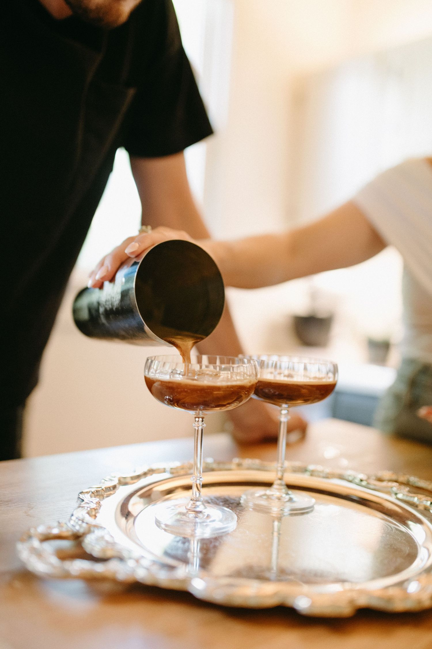 Cocktails being poured into coupe glasses on an ornate silver serving tray.