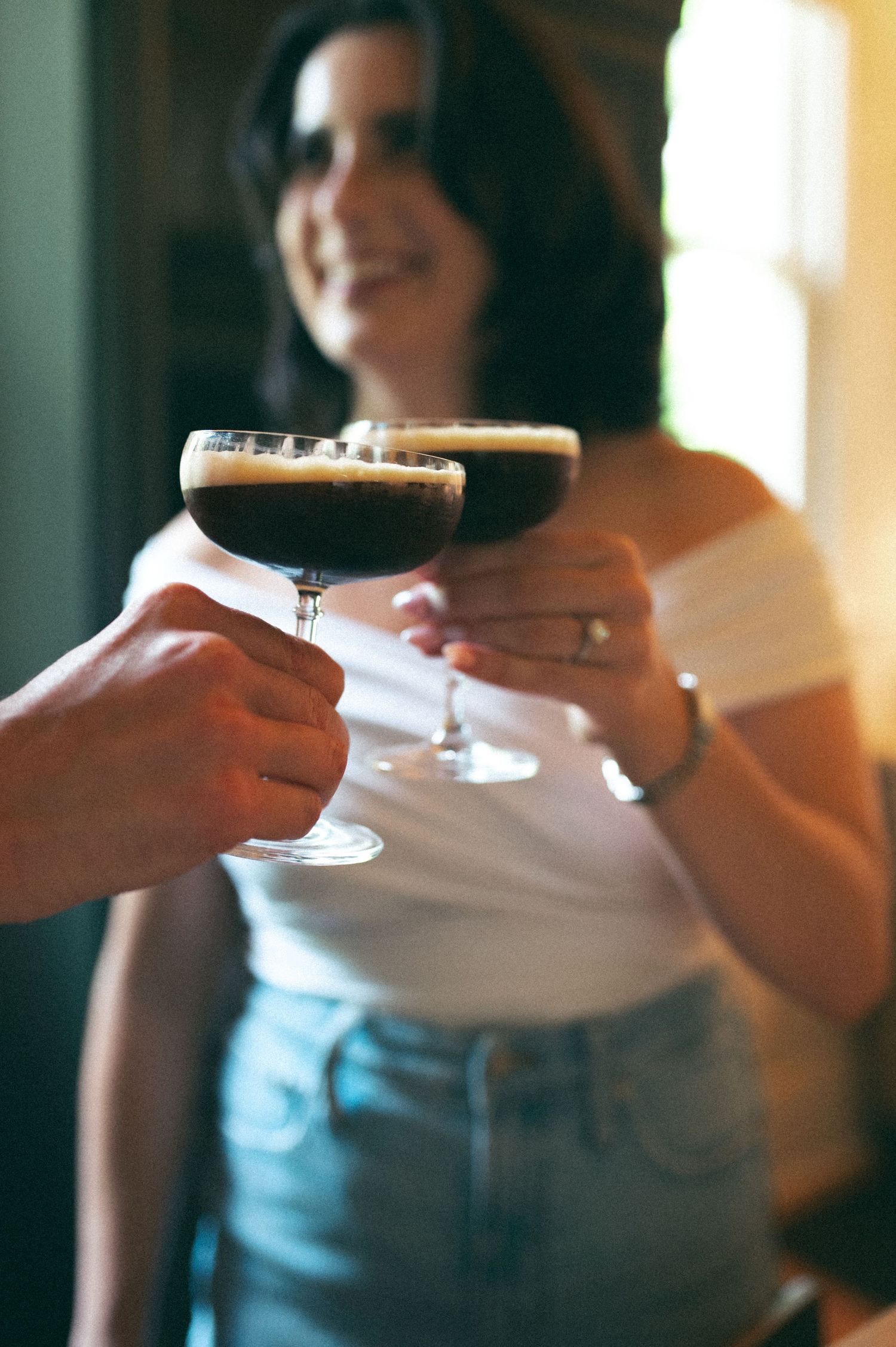 Two people toasting with dark cocktails in coupe glasses.