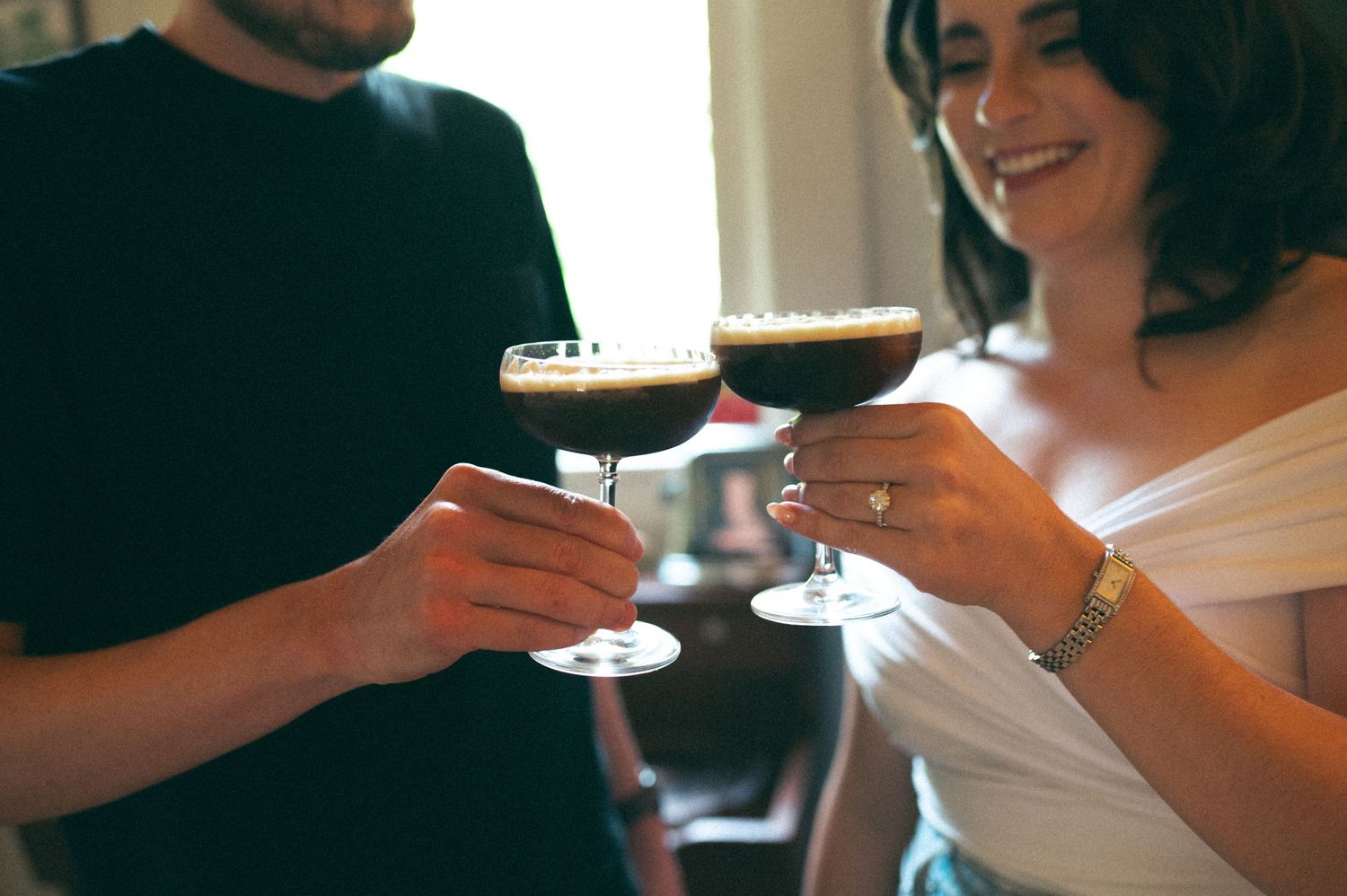 Two people clinking cocktail glasses in a sunlit kitchen.
