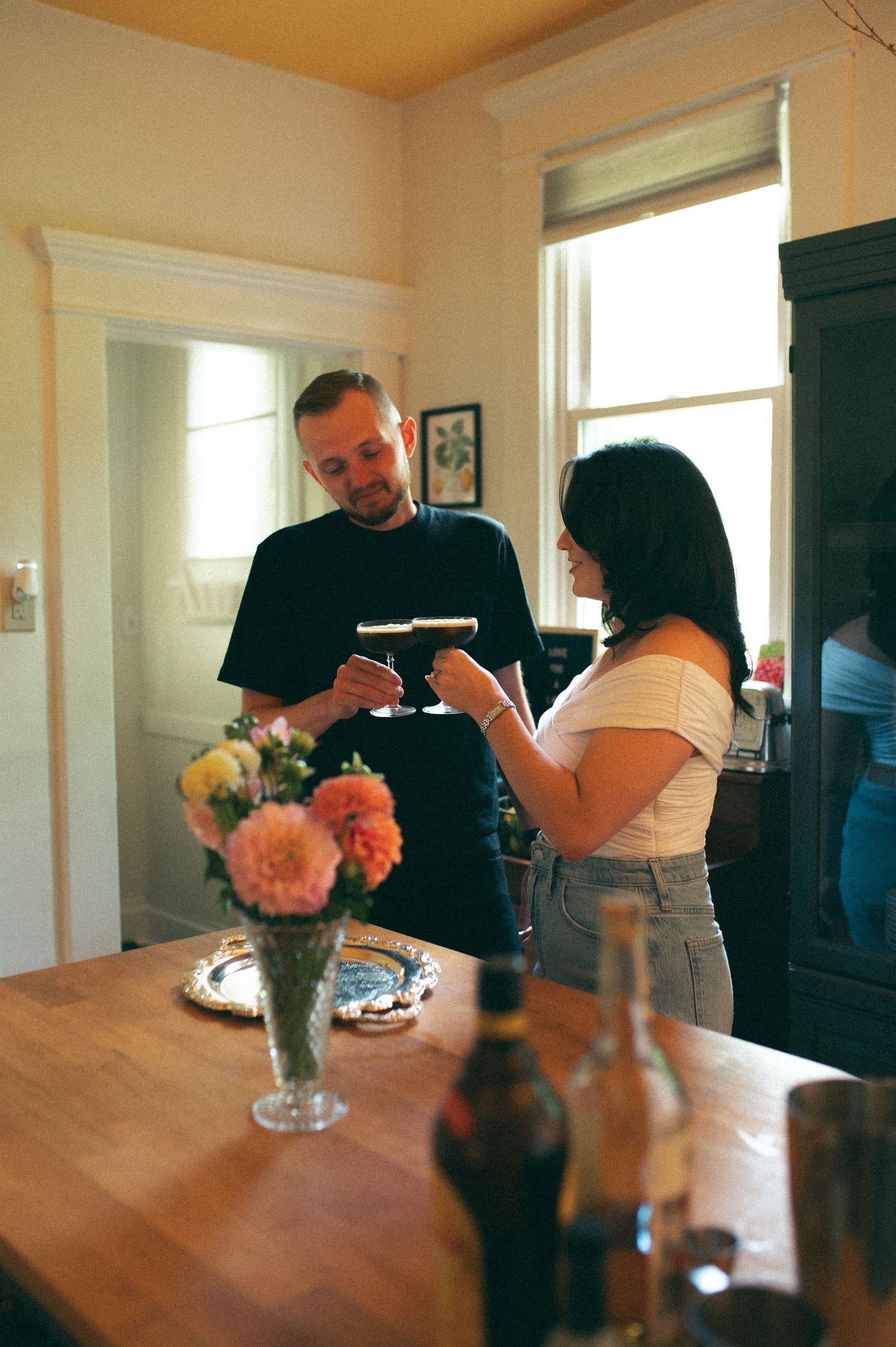 A couple sharing drinks at a kitchen counter with fresh flowers in a vase.