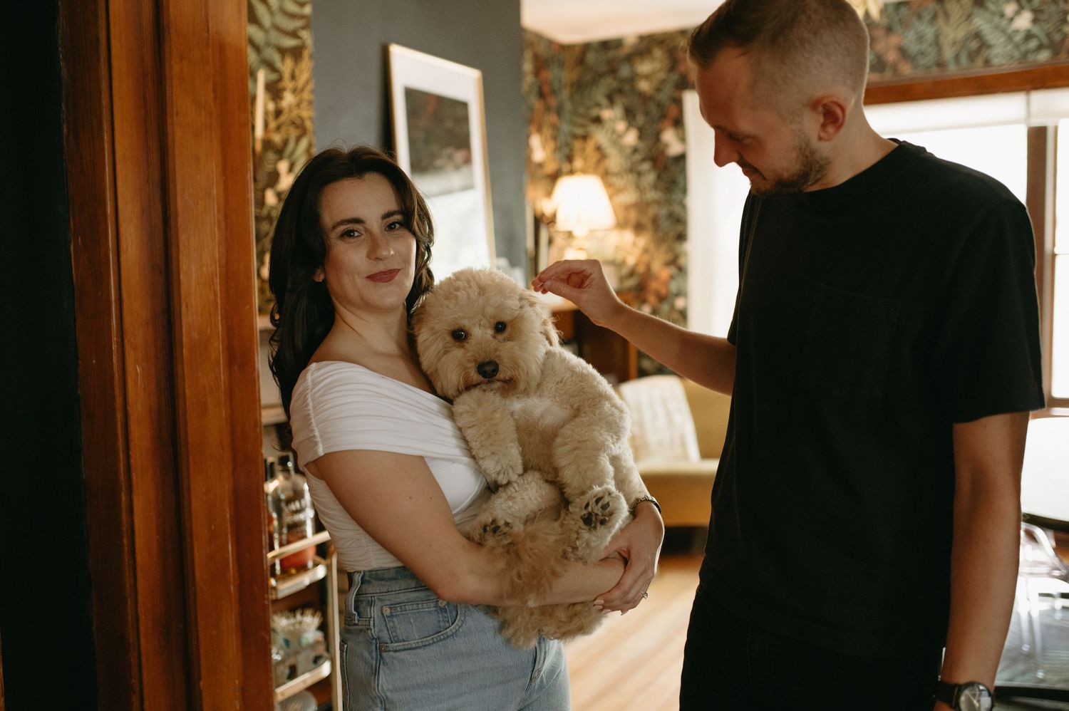 A couple standing in a cozy room while holding an adorable fluffy white dog.