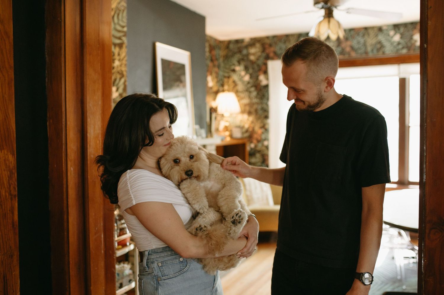A couple in casual attire sharing a tender moment while holding a small fluffy white dog indoors.