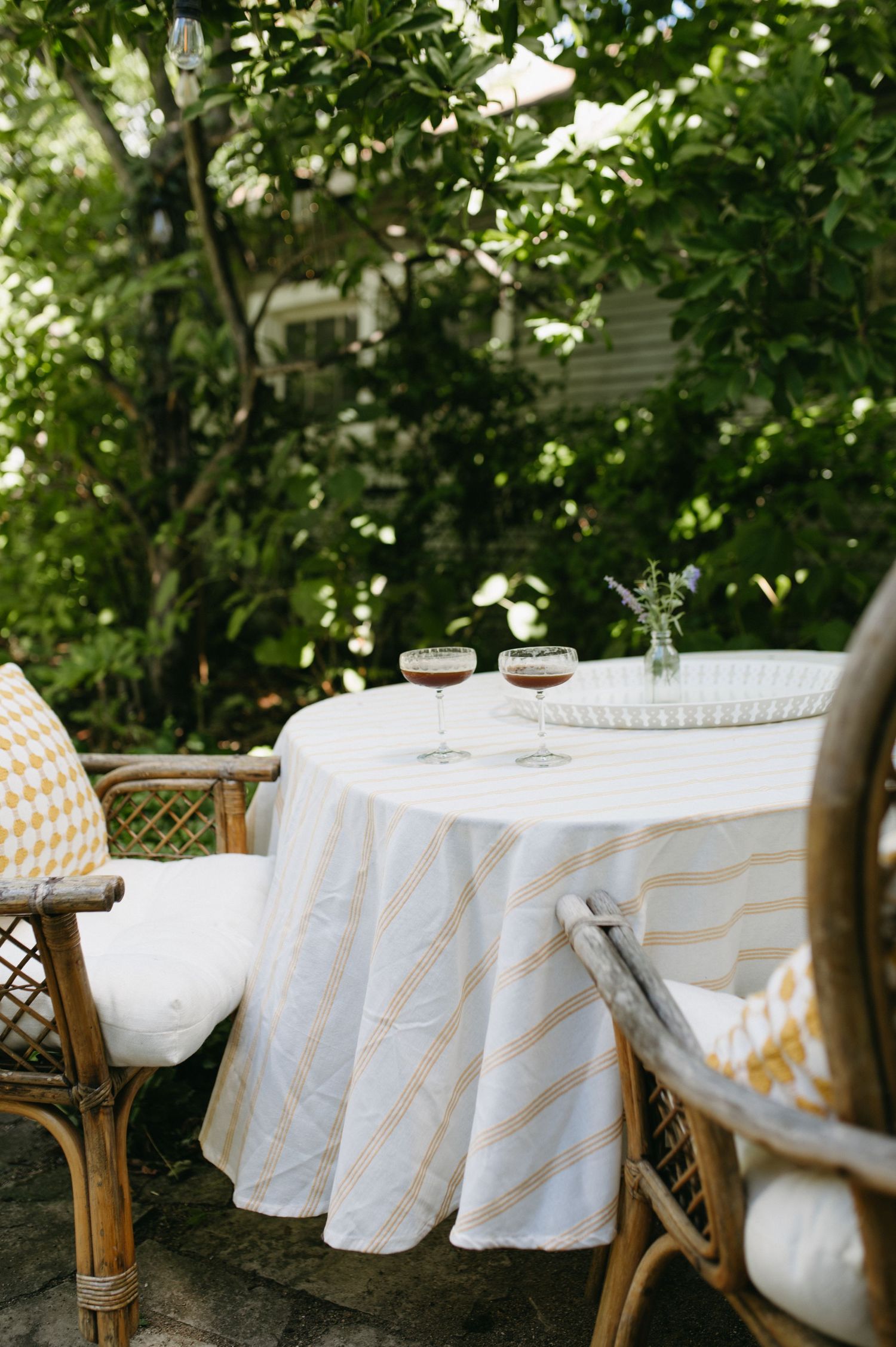 Outdoor garden dining setup with white tablecloth and rattan chairs under leafy trees.
