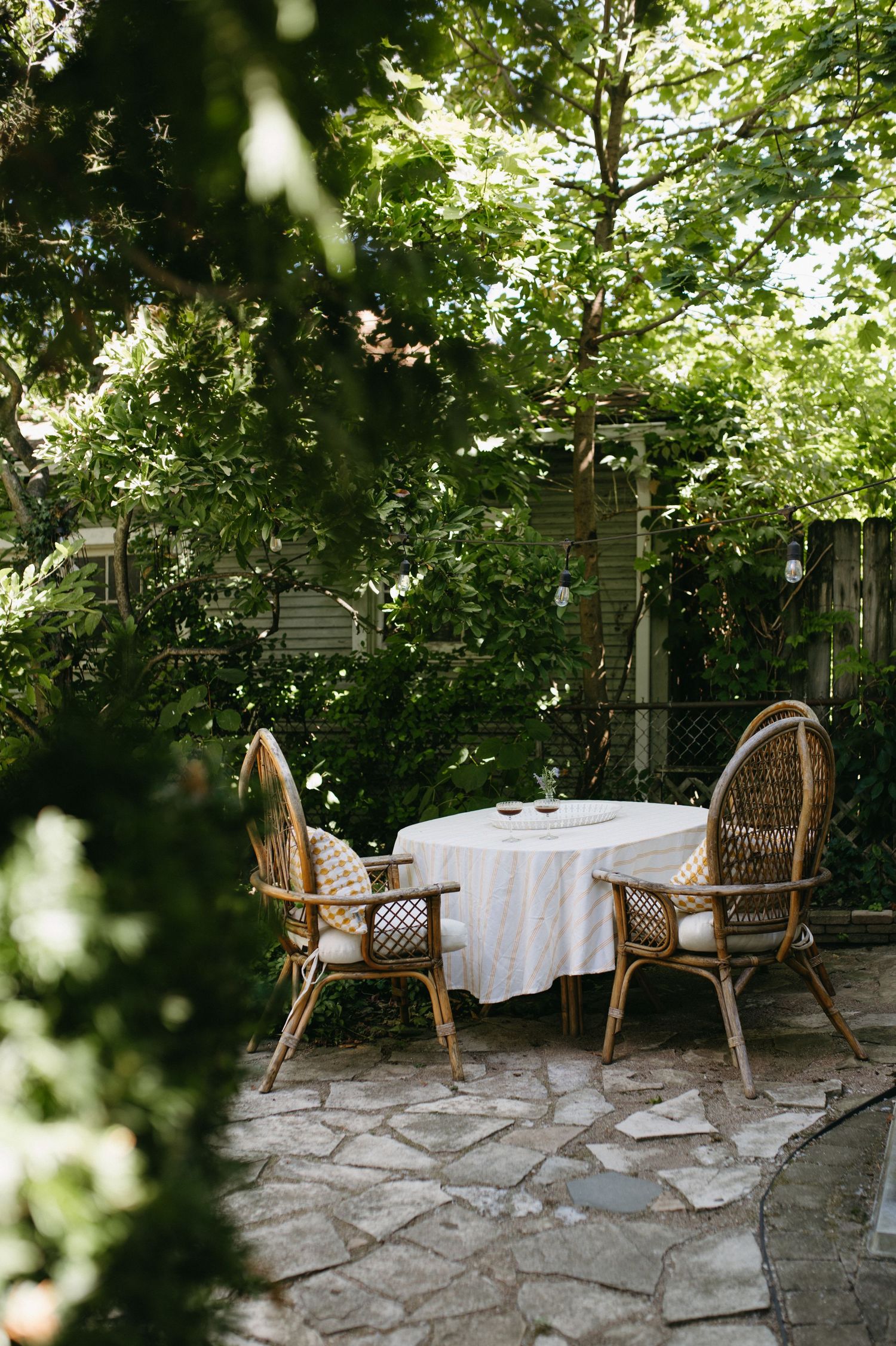 Intimate garden table setting with wicker chairs and stone patio surrounded by lush greenery.