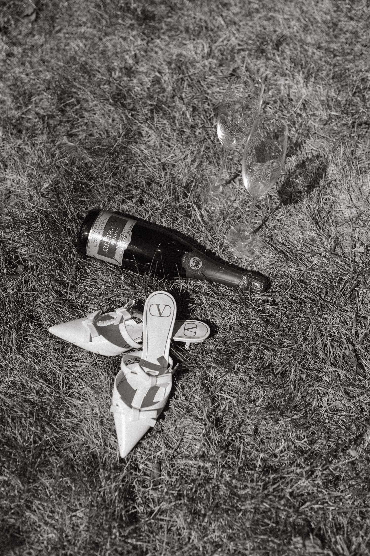 Black and white photo of a wine bottle and heels lying on grass.