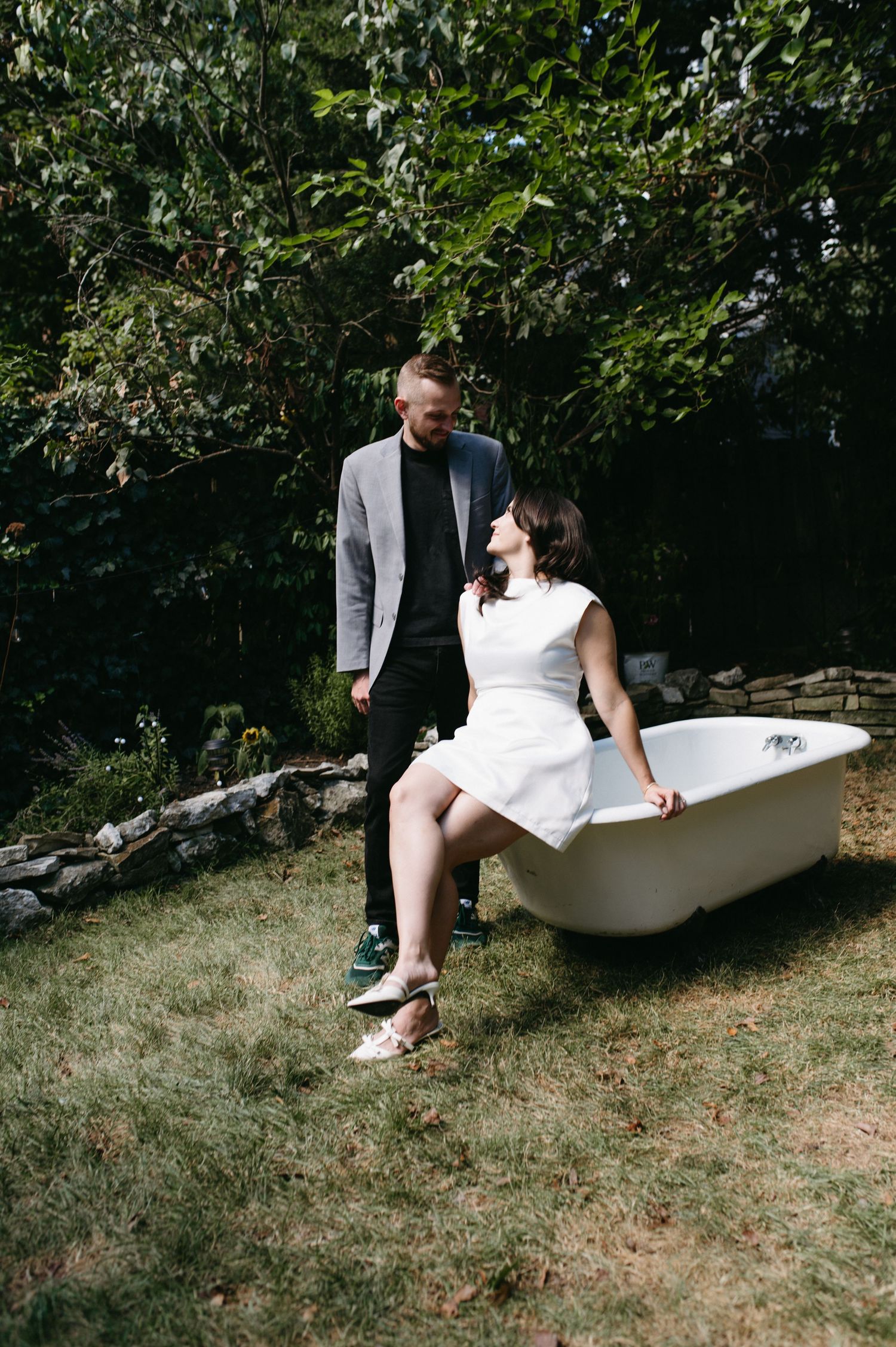 A couple sharing a tender moment near a white bathtub in an outdoor garden setting.