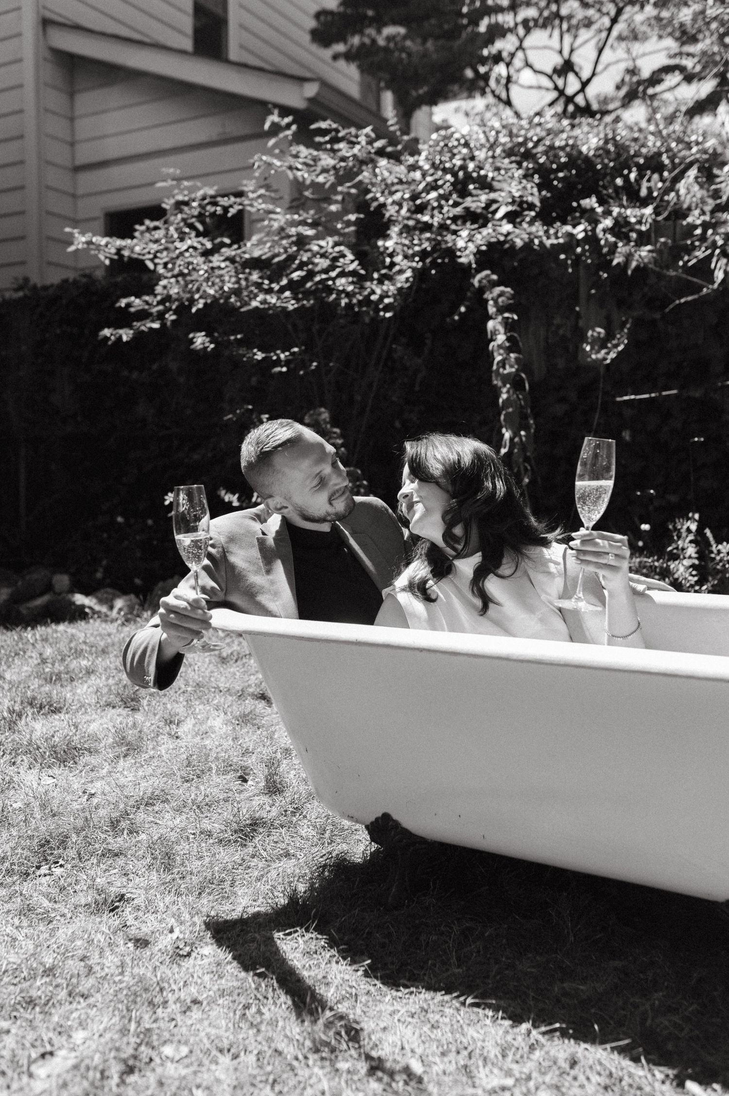 Black and white photo of couple sharing drinks in outdoor bathtub setup.