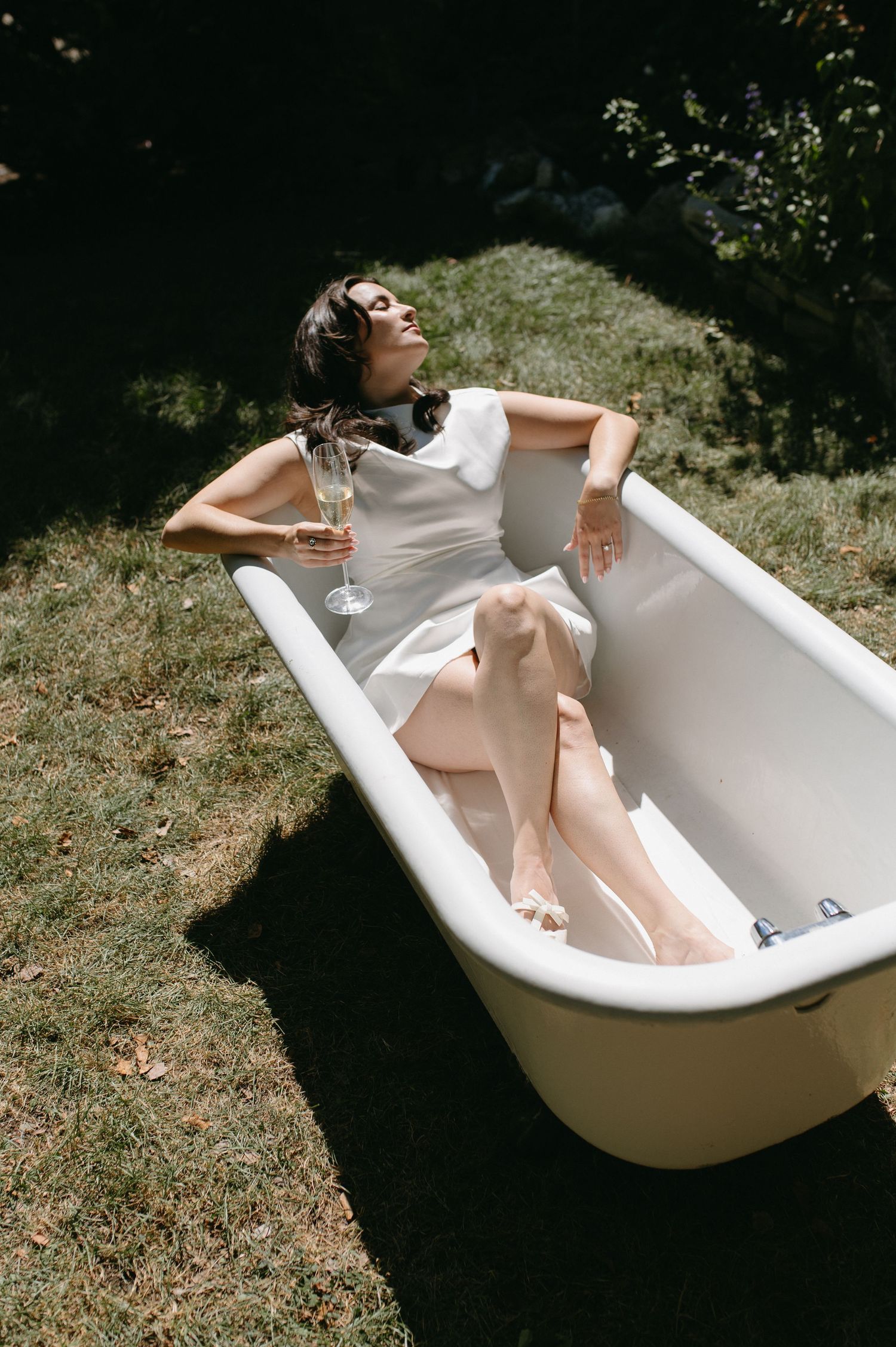 A carefree figure reclines in a vintage bathtub outdoors holding a wine glass while wearing a white dress.