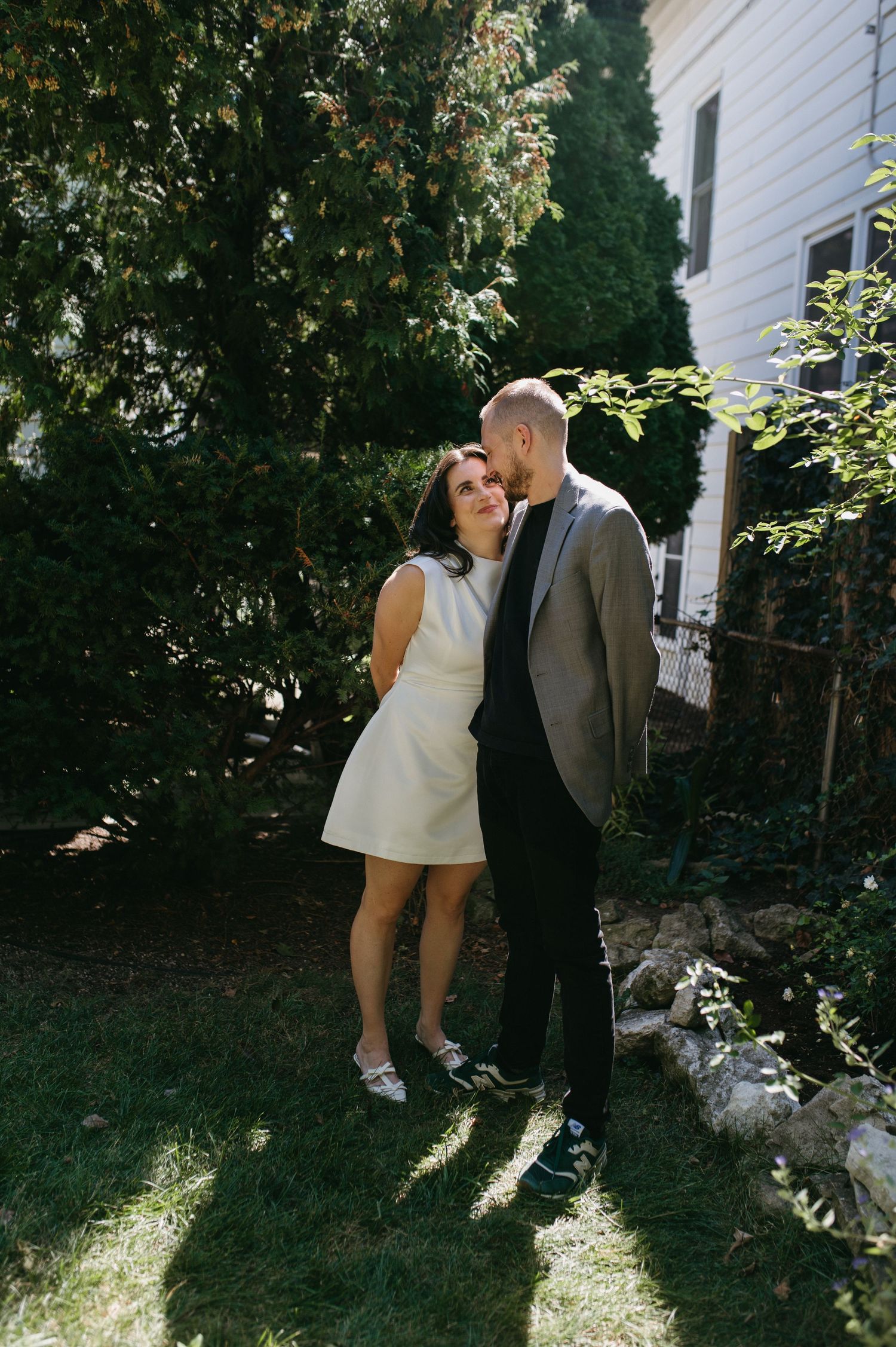 A couple embraces in a sunny garden setting beside a white house.
