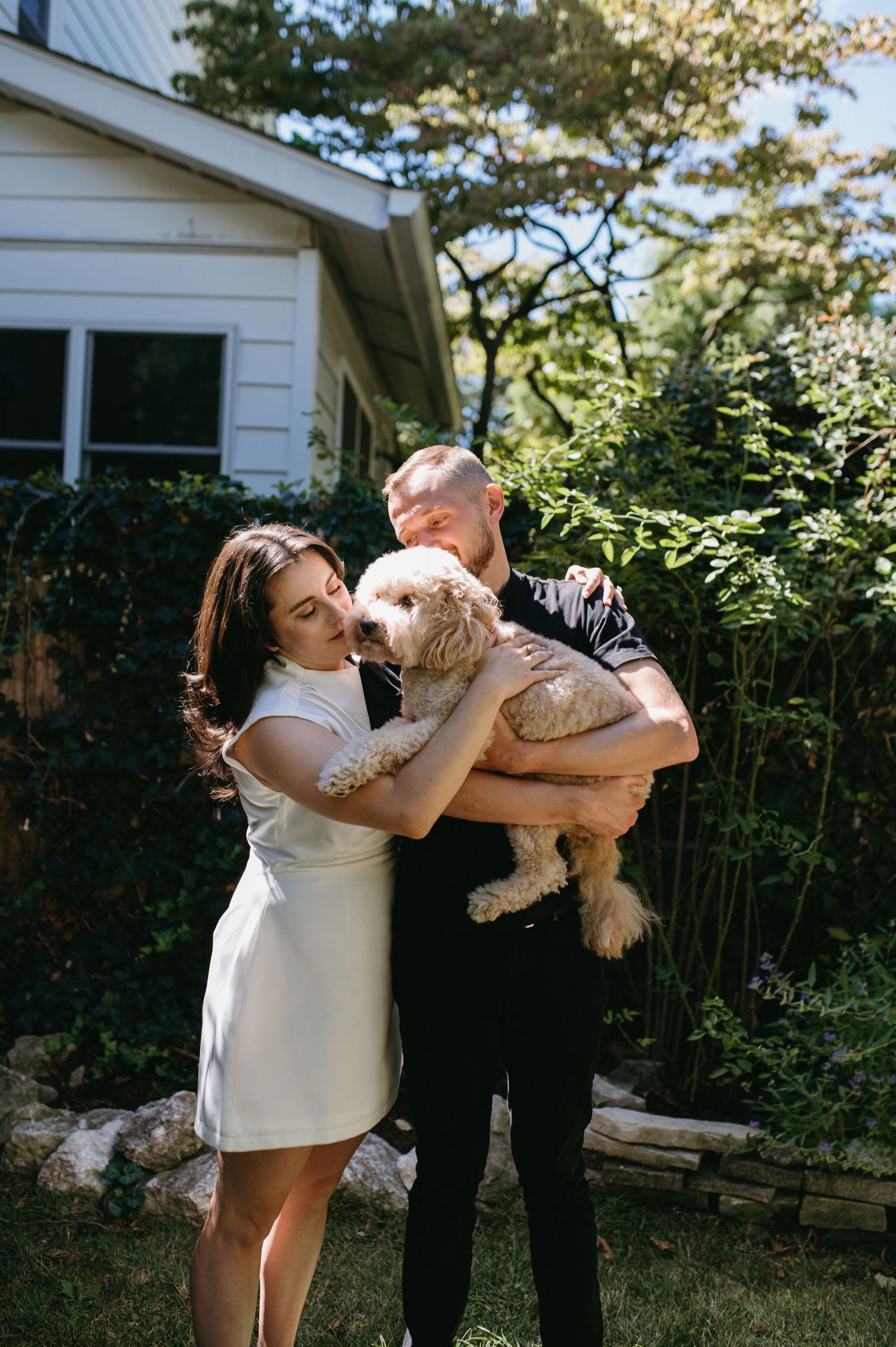 A couple holds their beloved goldendoodle dog while standing in their backyard garden.