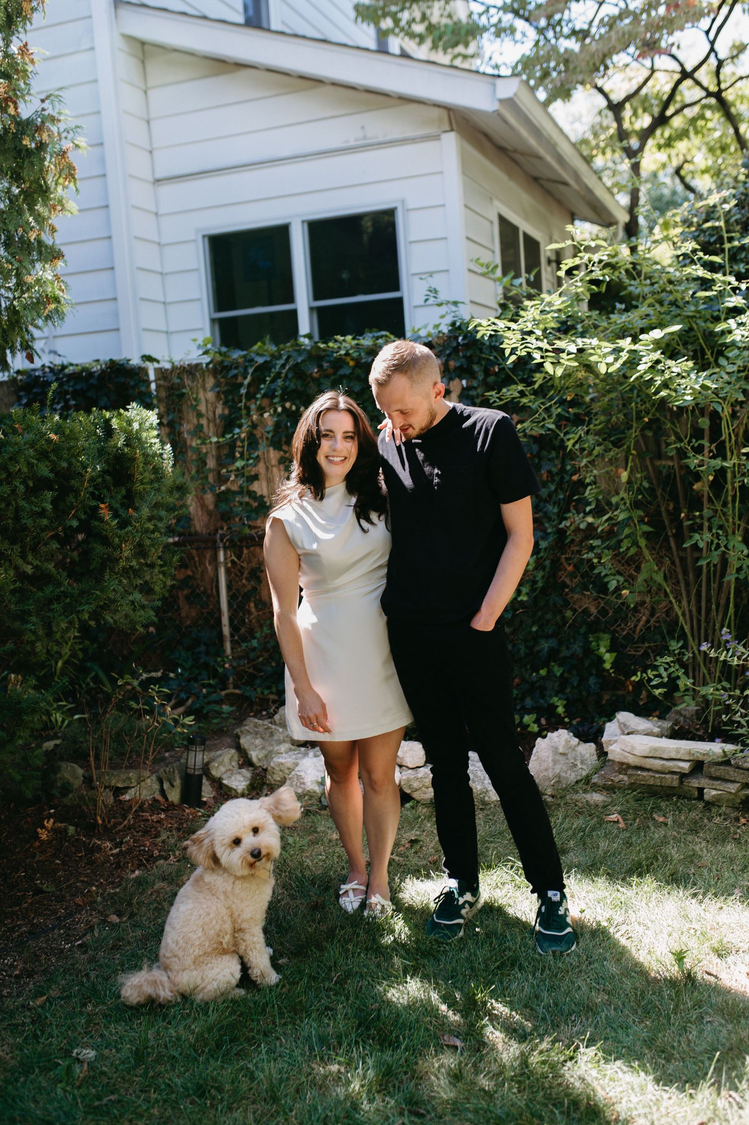 A couple poses with their goldendoodle in front of a white house on a sunny day.