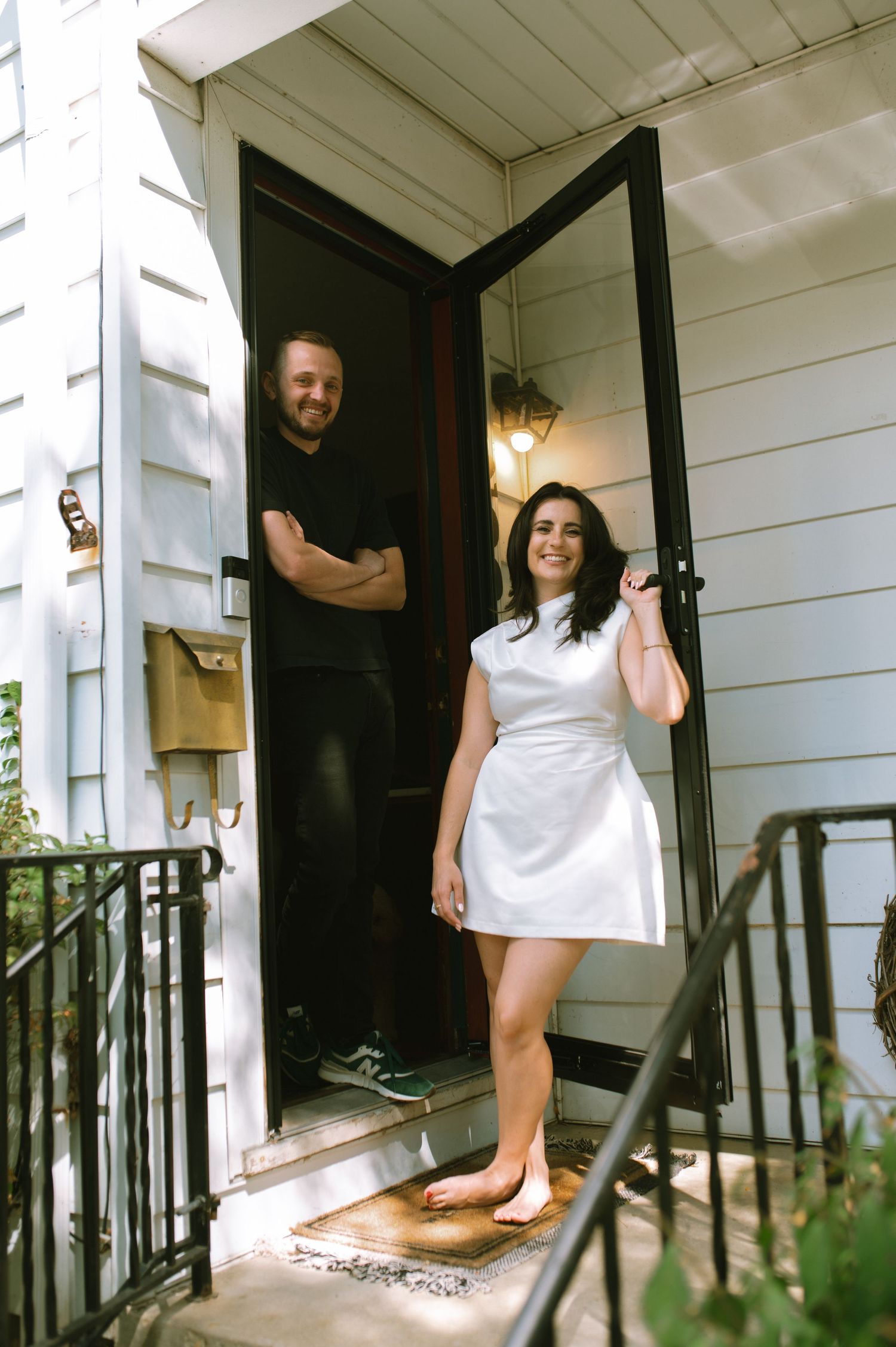 A couple stands in their doorway entrance of a white house on a sunny day.