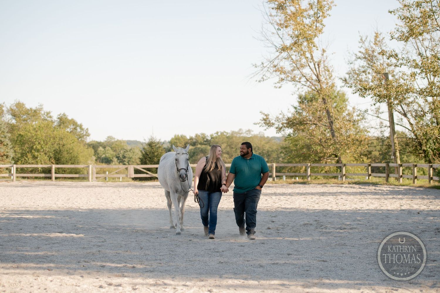 A grey horse follows two people walking through a sandy arena.