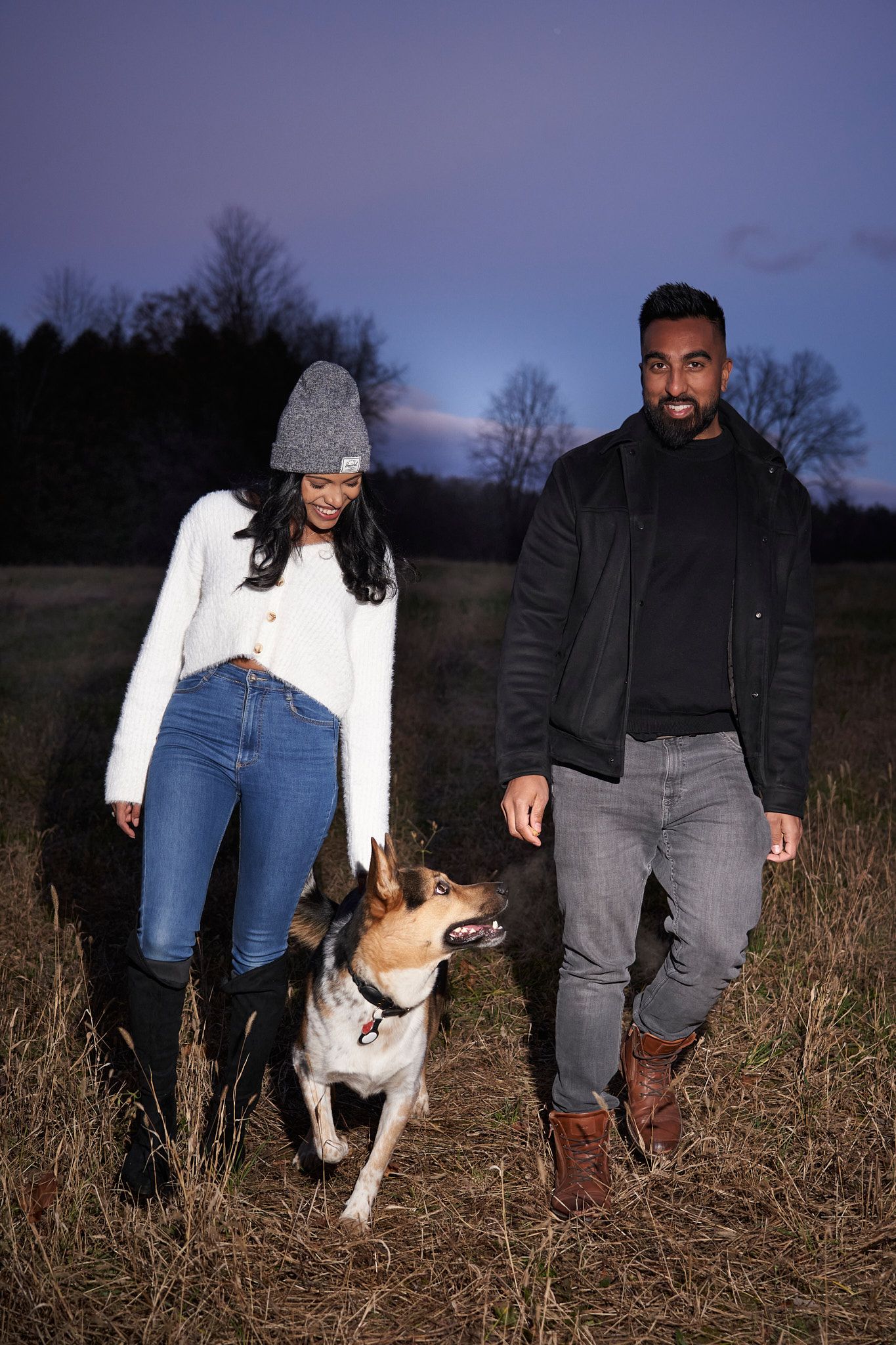 A couple walking their brown and white dog through a field at dusk with the woman wearing a beanie and white sweater.