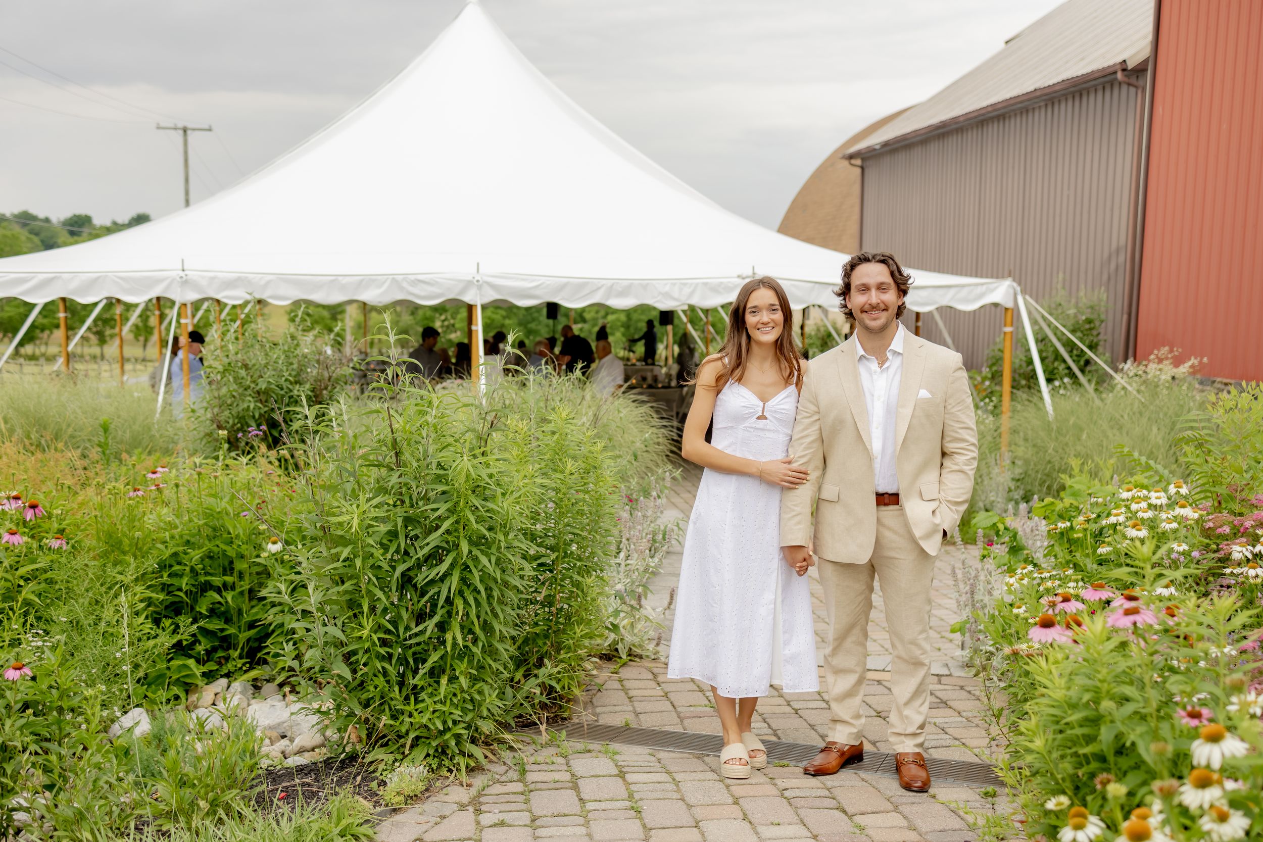 Couple in wedding attire standing on brick path surrounded by wildflowers near white tent and red barn.