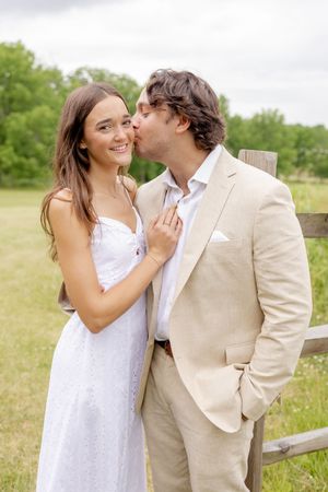 A couple in wedding attire shares a romantic kiss outdoors near a wooden fence in a green field.