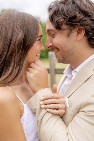 Smiling couple in romantic close embrace, woman wearing engagement ring, man in beige suit, outdoors with greenery background.