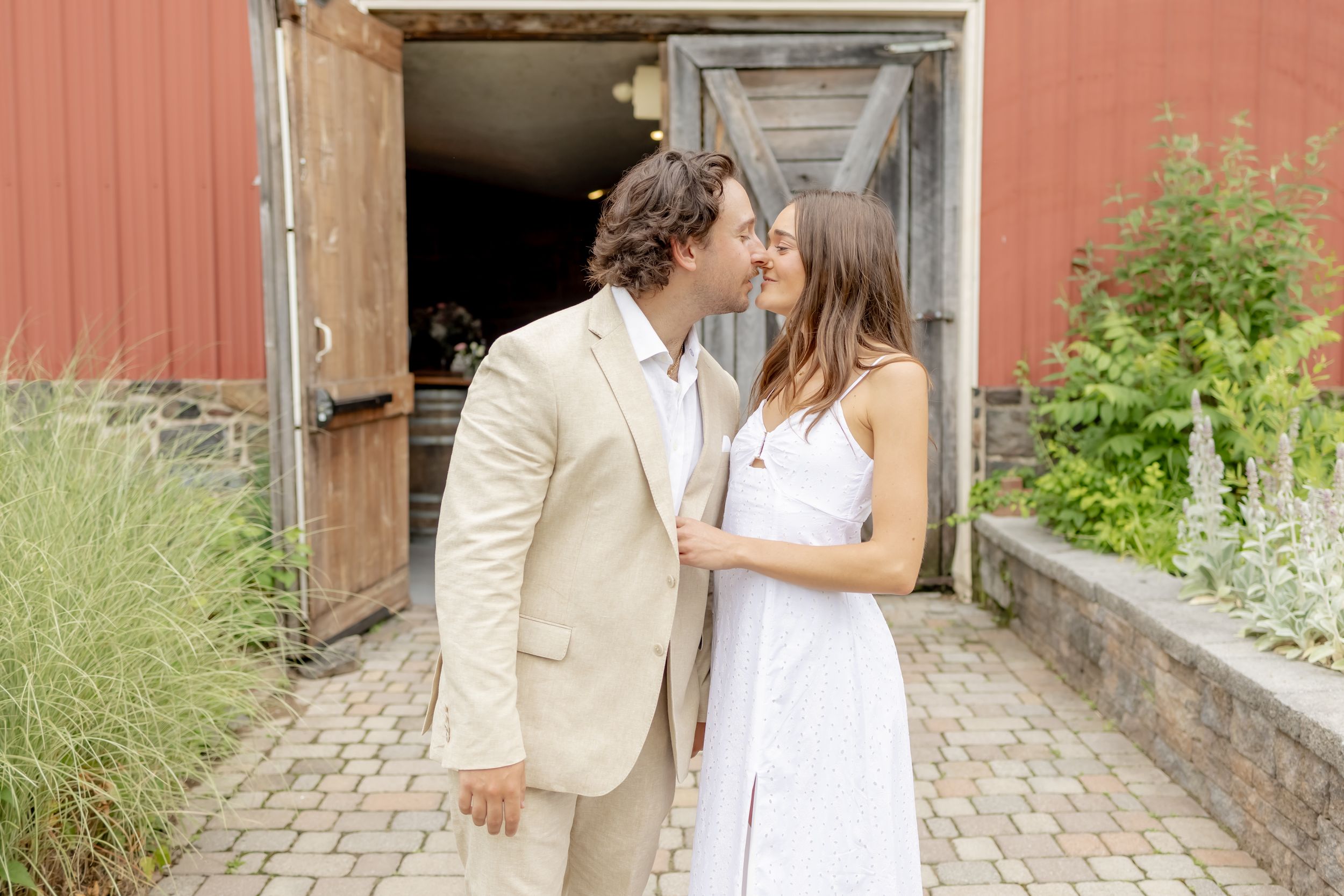 Couple sharing a romantic kiss outside a rustic red barn, man in beige suit and woman in white dress.