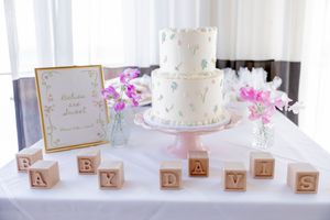 Two-tier white baby shower cake on pink stand with wooden letter blocks in Long Branch, NJ.