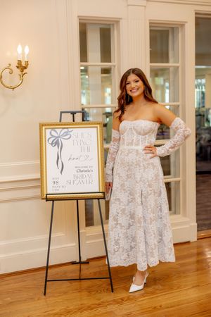 Smiling bride in white lace midi dress posing beside bridal shower welcome sign.