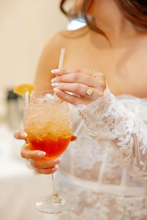 Bride in white lace dress holding a large orange cocktail with lemon garnish and straw.