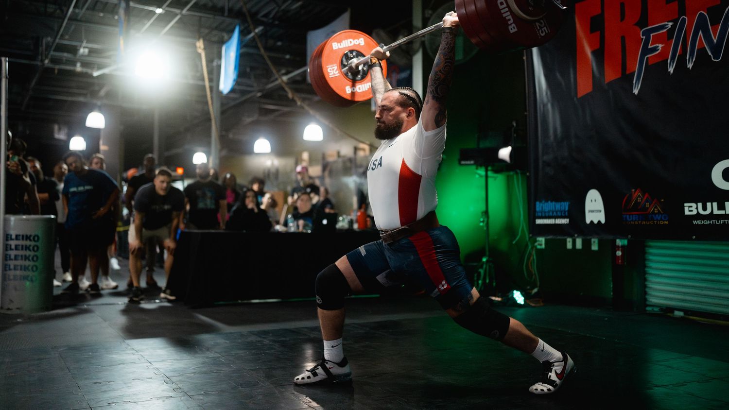 Weightlifter performing a snatch lift at a dimly lit indoor gym competition with spectators watching.