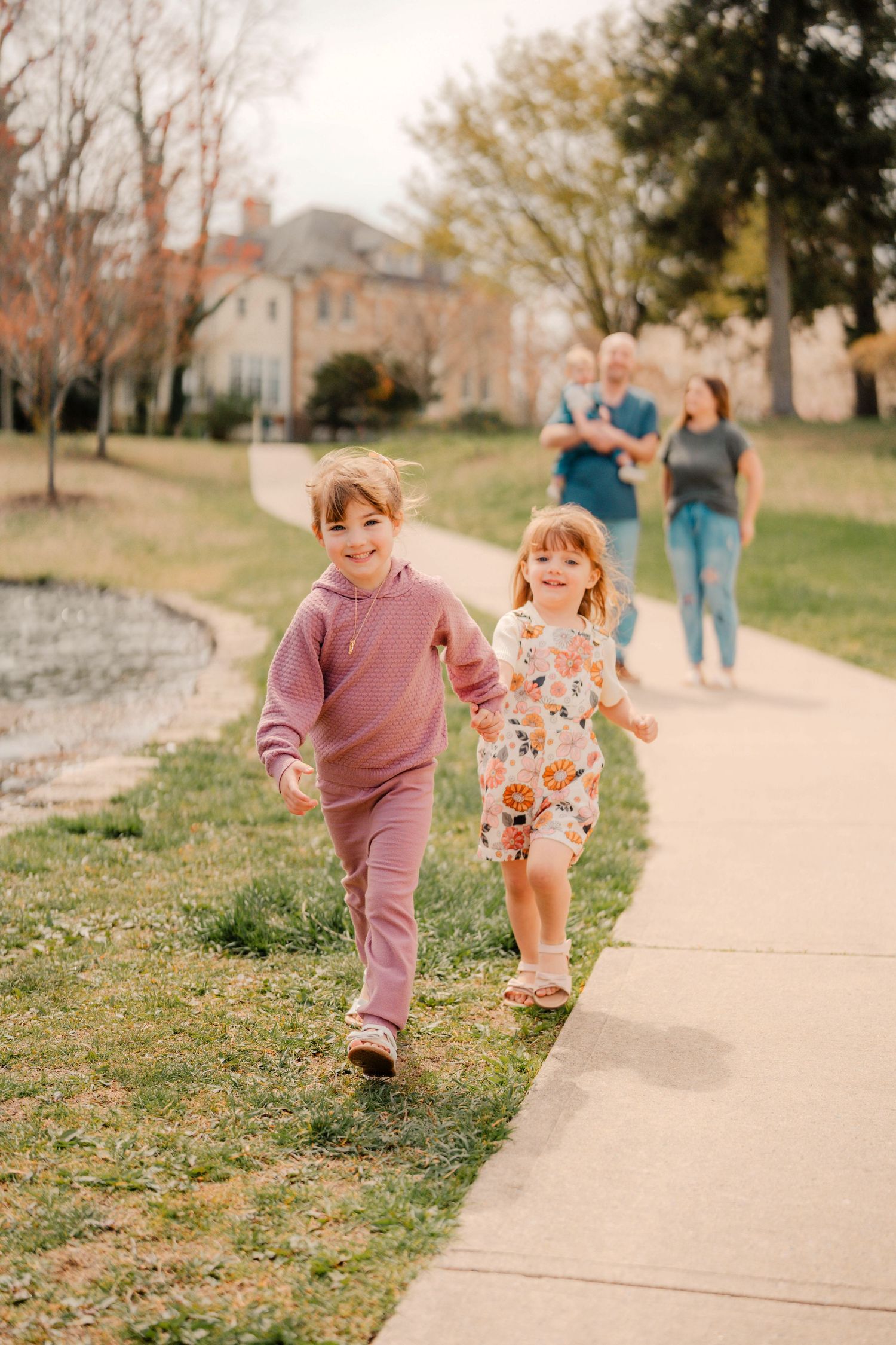 Two young children in coordinating pink and floral outfits run playfully along a concrete path on a sunny autumn day.
