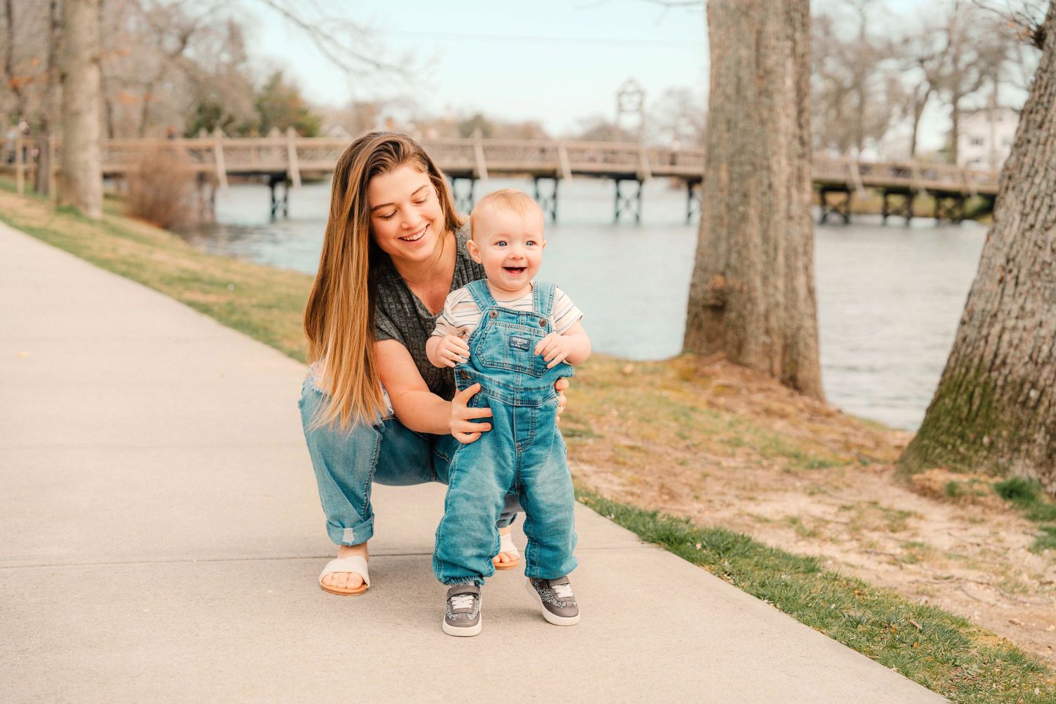 A toddler in denim overalls smiles while standing on a pathway near a lake with a wooden bridge in the background.